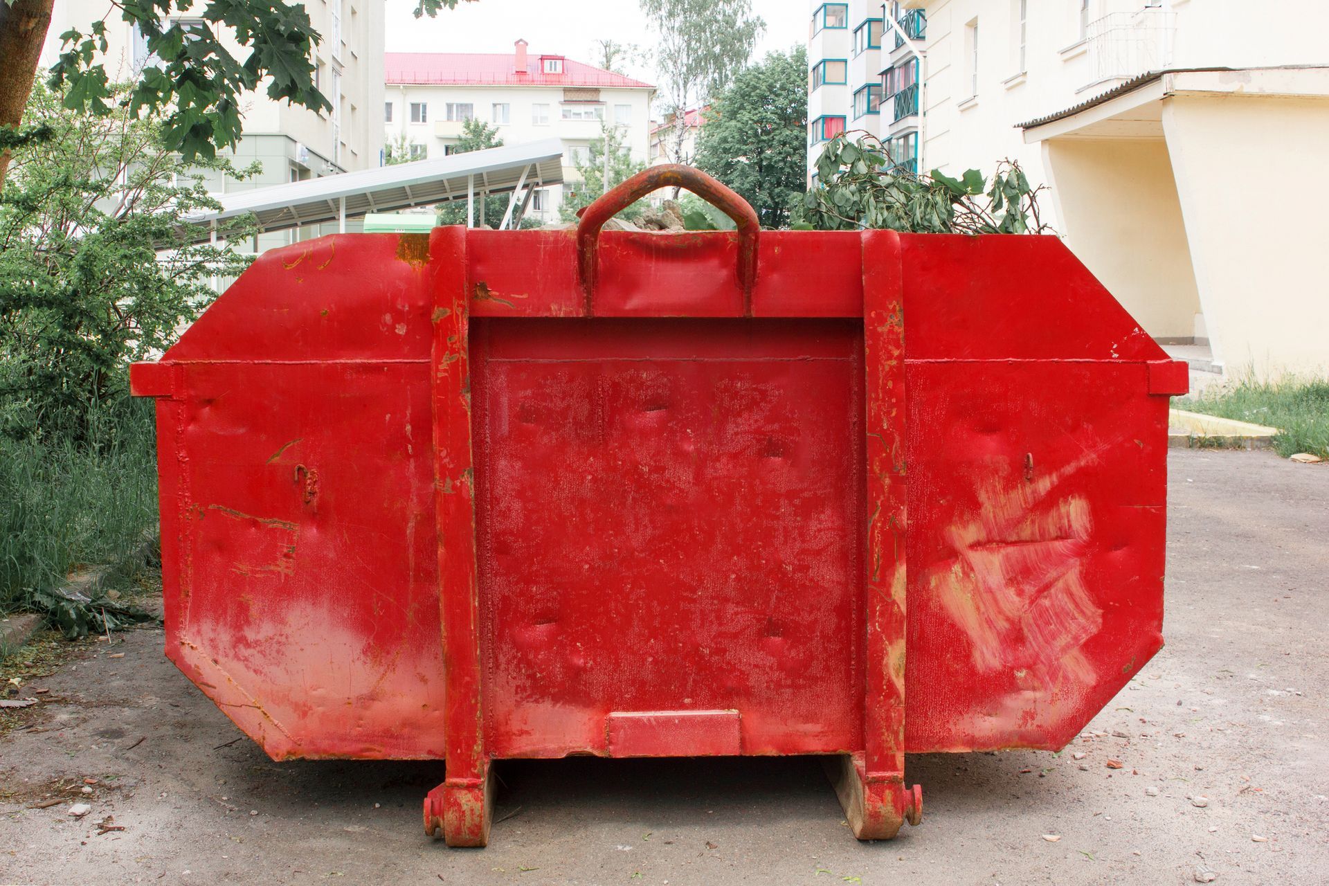 Red dumpster on a paved surface, against a backdrop of buildings and greenery.