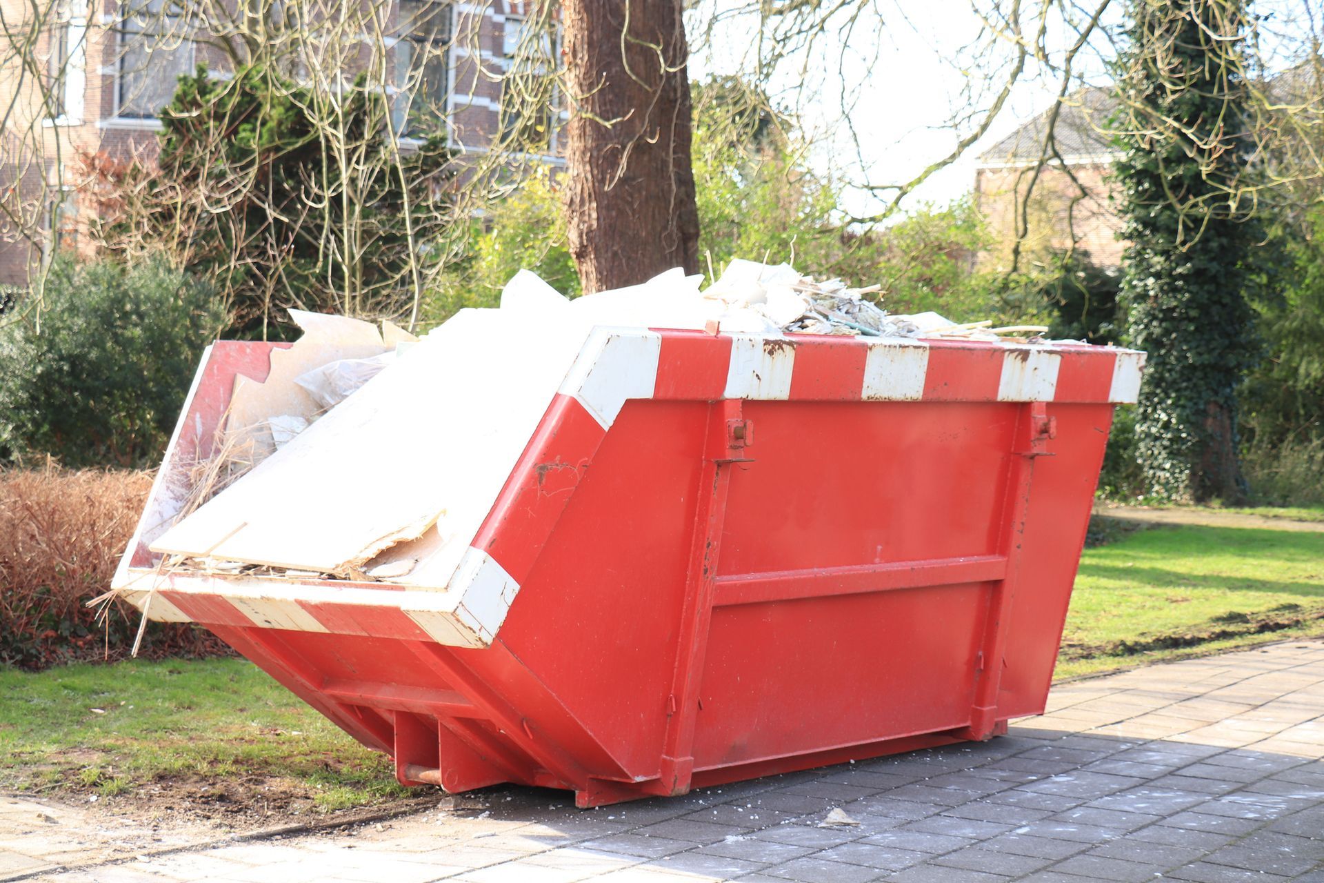 Red dumpster filled with debris on a paved surface near a tree and greenery.