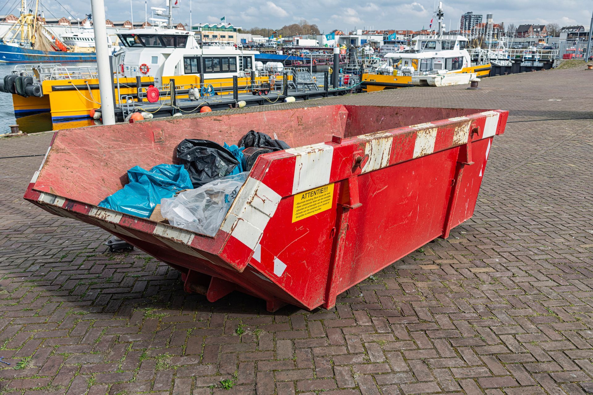 Red dumpster on a brick surface, filled with garbage, with boats in the background.
