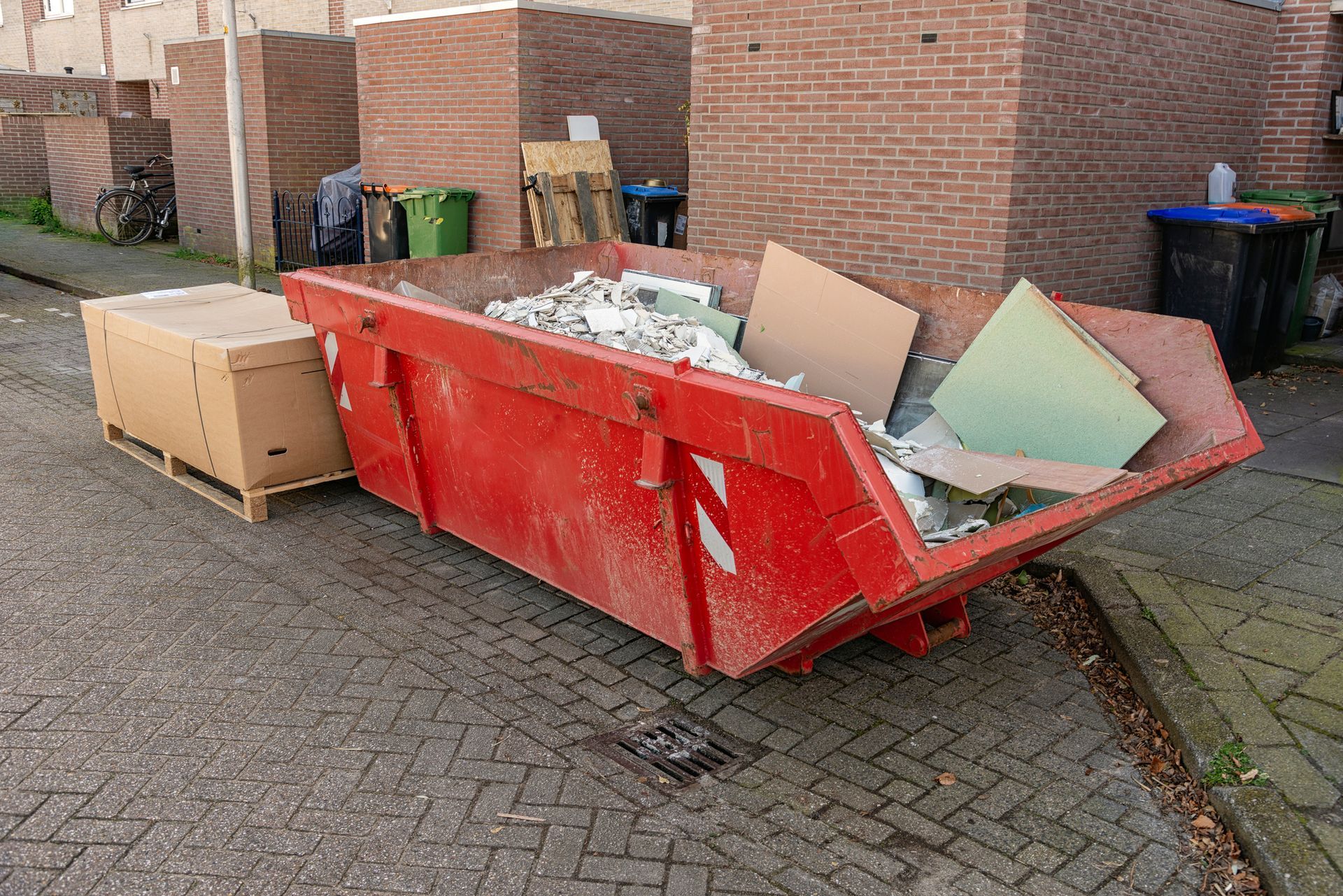 Red dumpster filled with construction debris on a city street, cardboard box beside it.