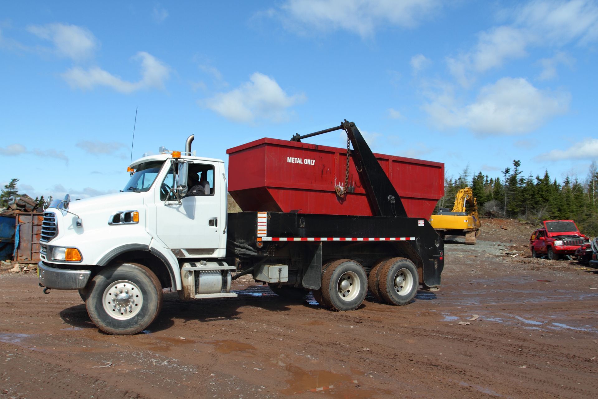 White truck with red container on a brown lot, cloudy sky, trees in background, and construction equipment nearby.