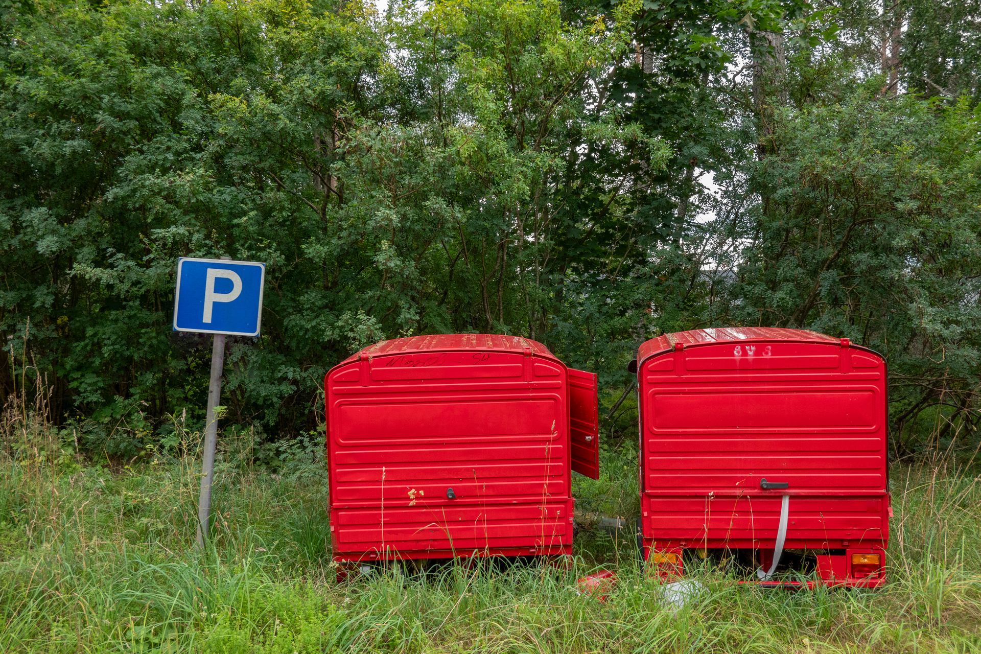 Two red trailers parked next to a parking sign with a backdrop of green trees.
