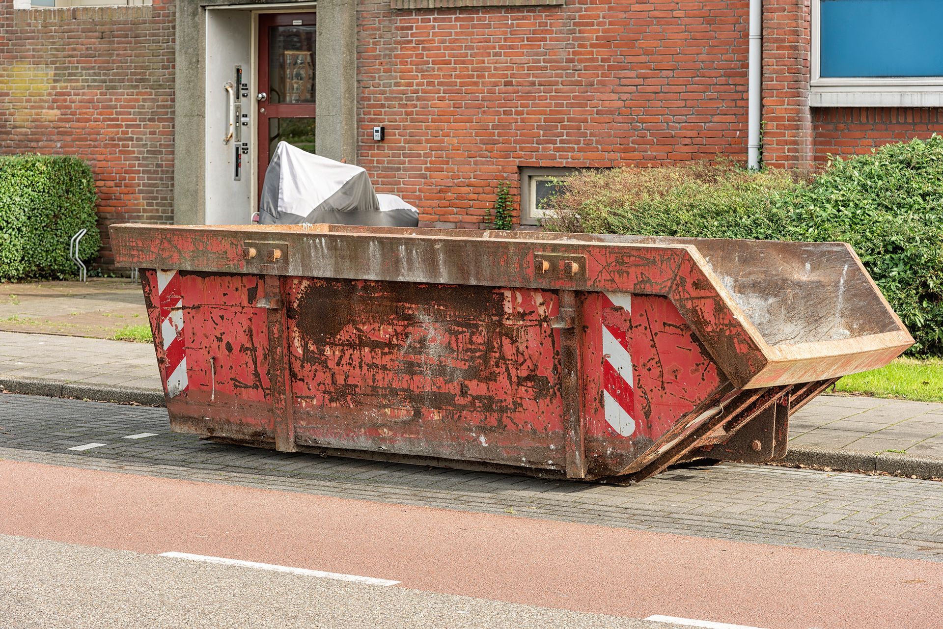 Red dumpster on a sidewalk next to a brick building.