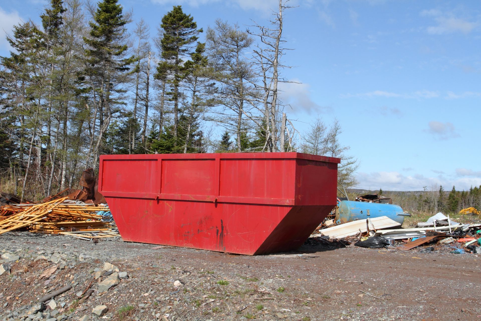 Red dumpster in a gravel lot surrounded by trees and debris on a sunny day.