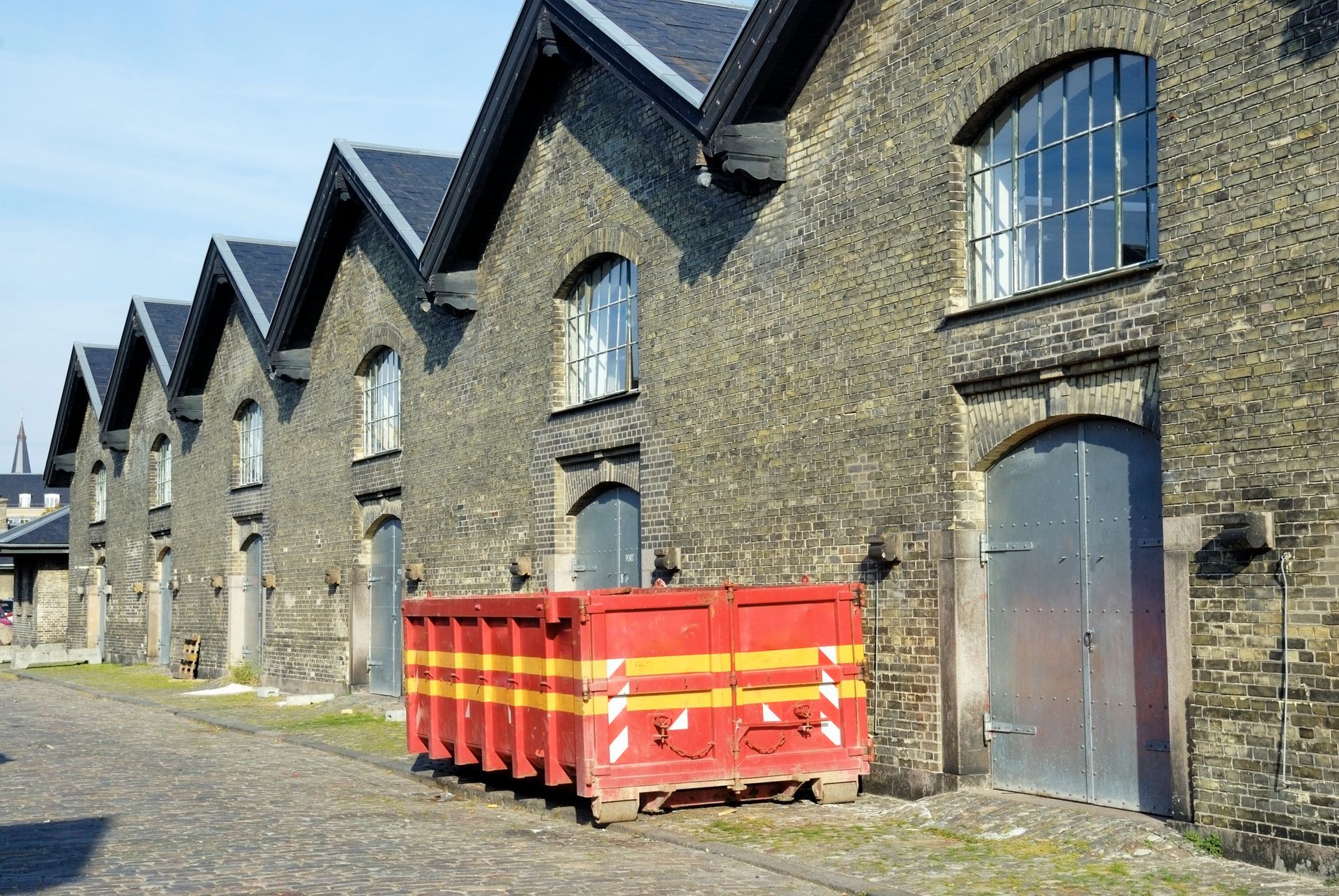 Row of old brick buildings with gable roofs. Red waste container in front.