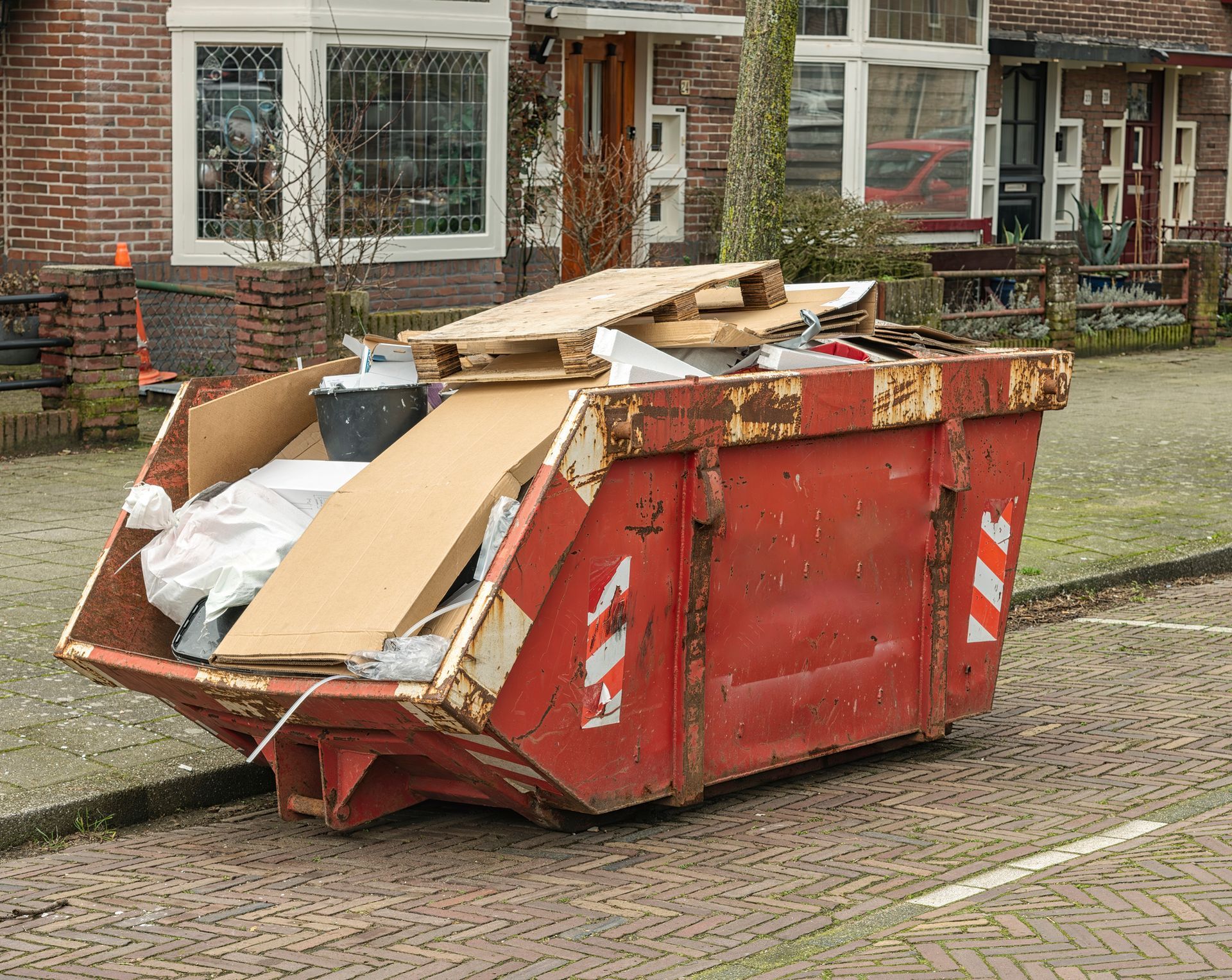 Red dumpster overflowing with trash on a brick street; houses in background.