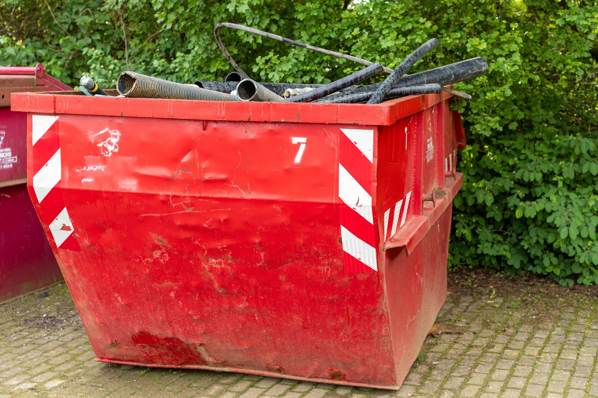 Red dumpster, partially filled with construction debris, with white diagonal stripes, outdoors.