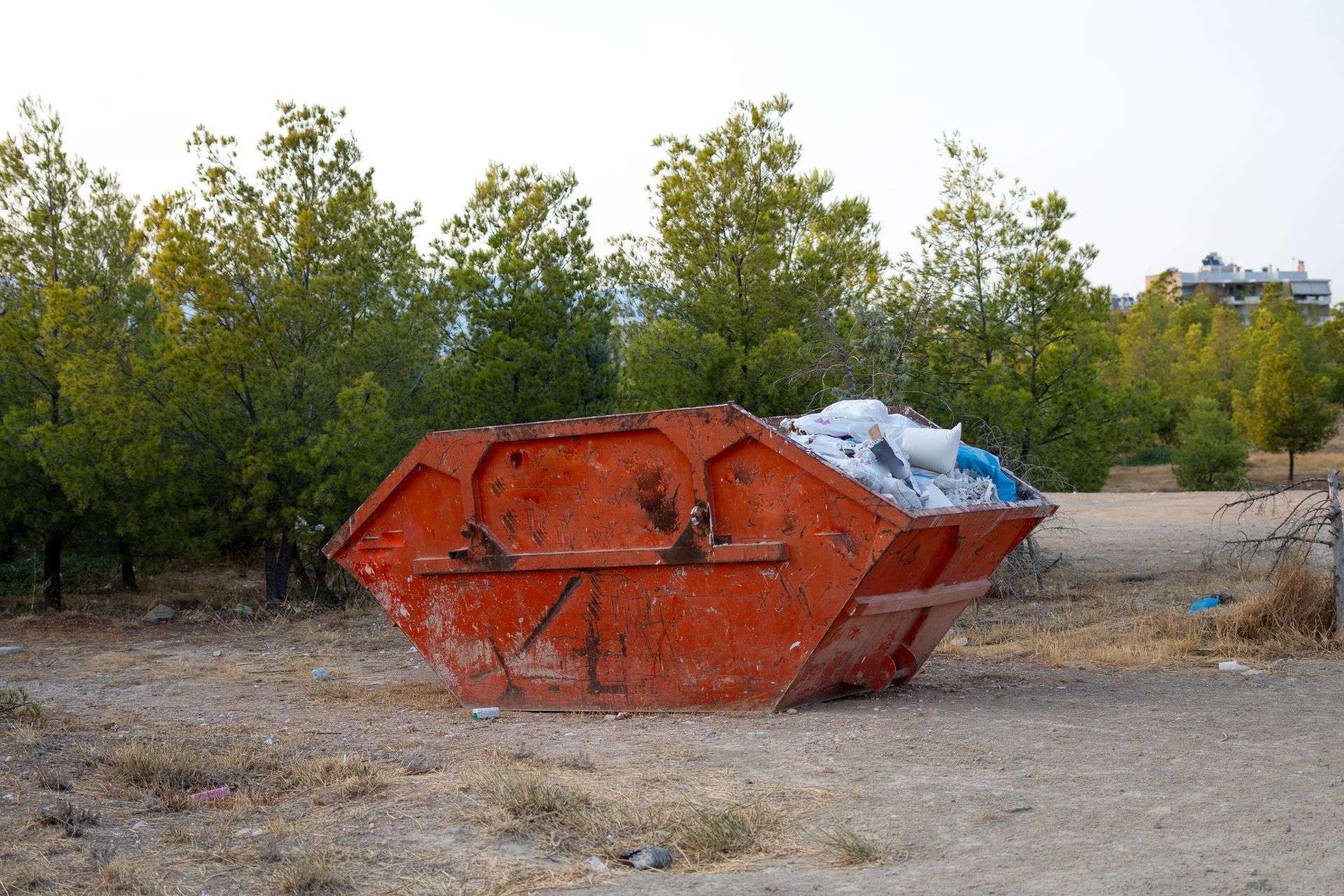 Orange dumpster filled with trash on a gravel lot, trees in the background.