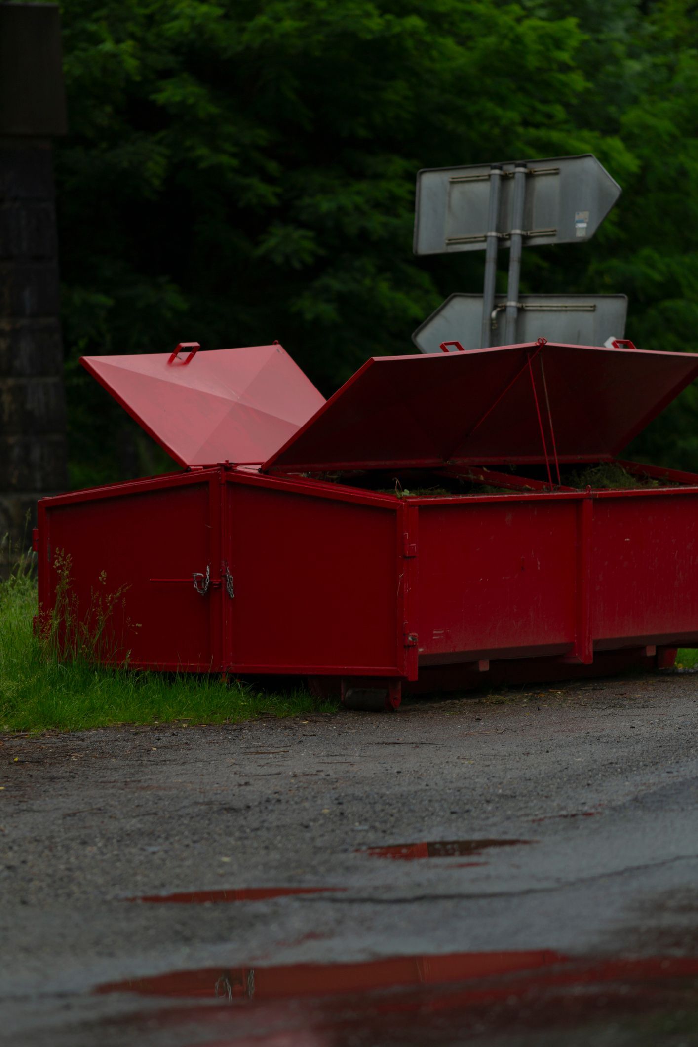 Red metal box with open lids beside a road, with signs and green foliage in the background.