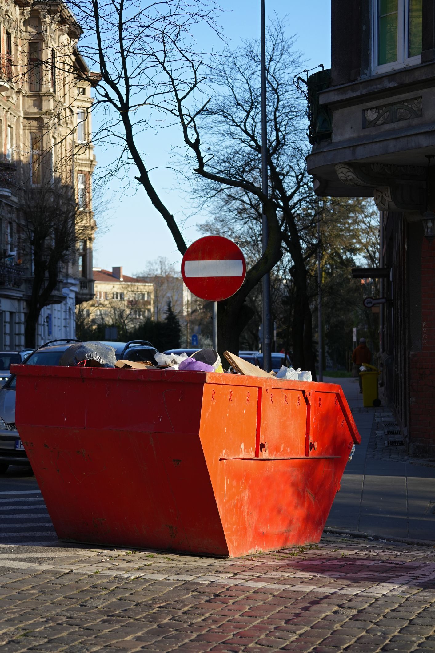 Bright red dumpster blocking a sidewalk on a city street, with a 