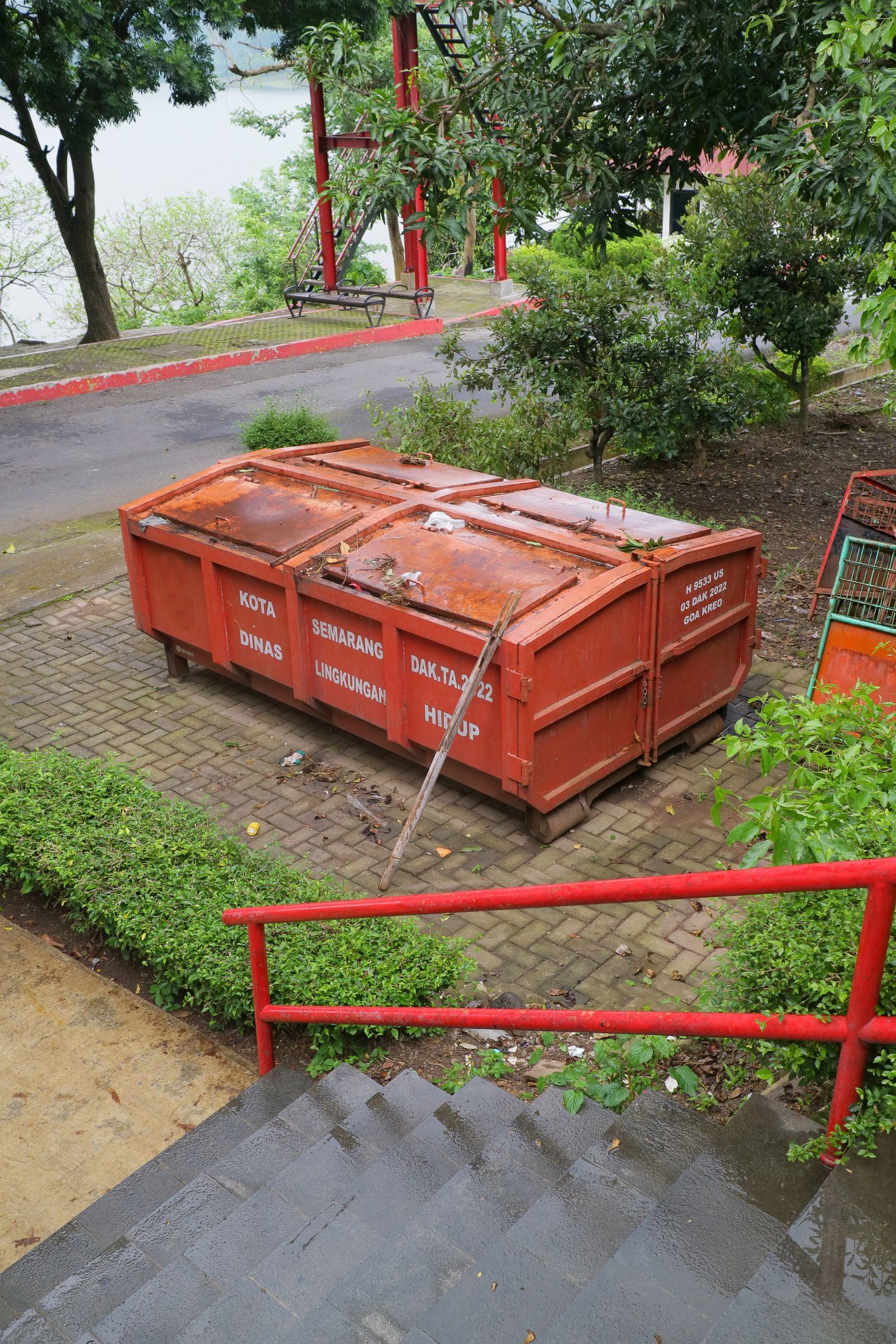 Red dumpster on concrete, near stairs and greenery, with a lake in the background.