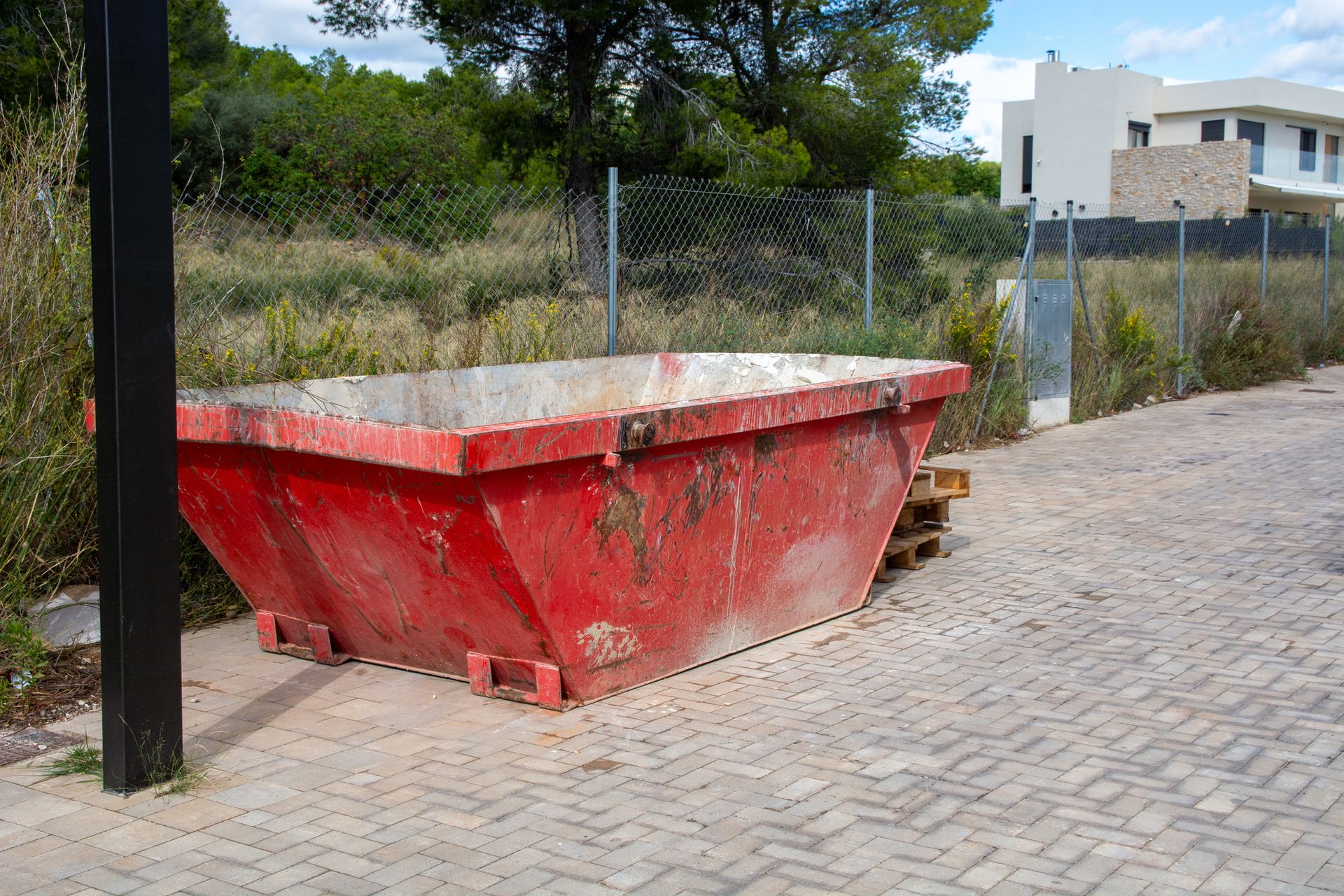Red dumpster on a paved area with a metal fence and vegetation in the background.