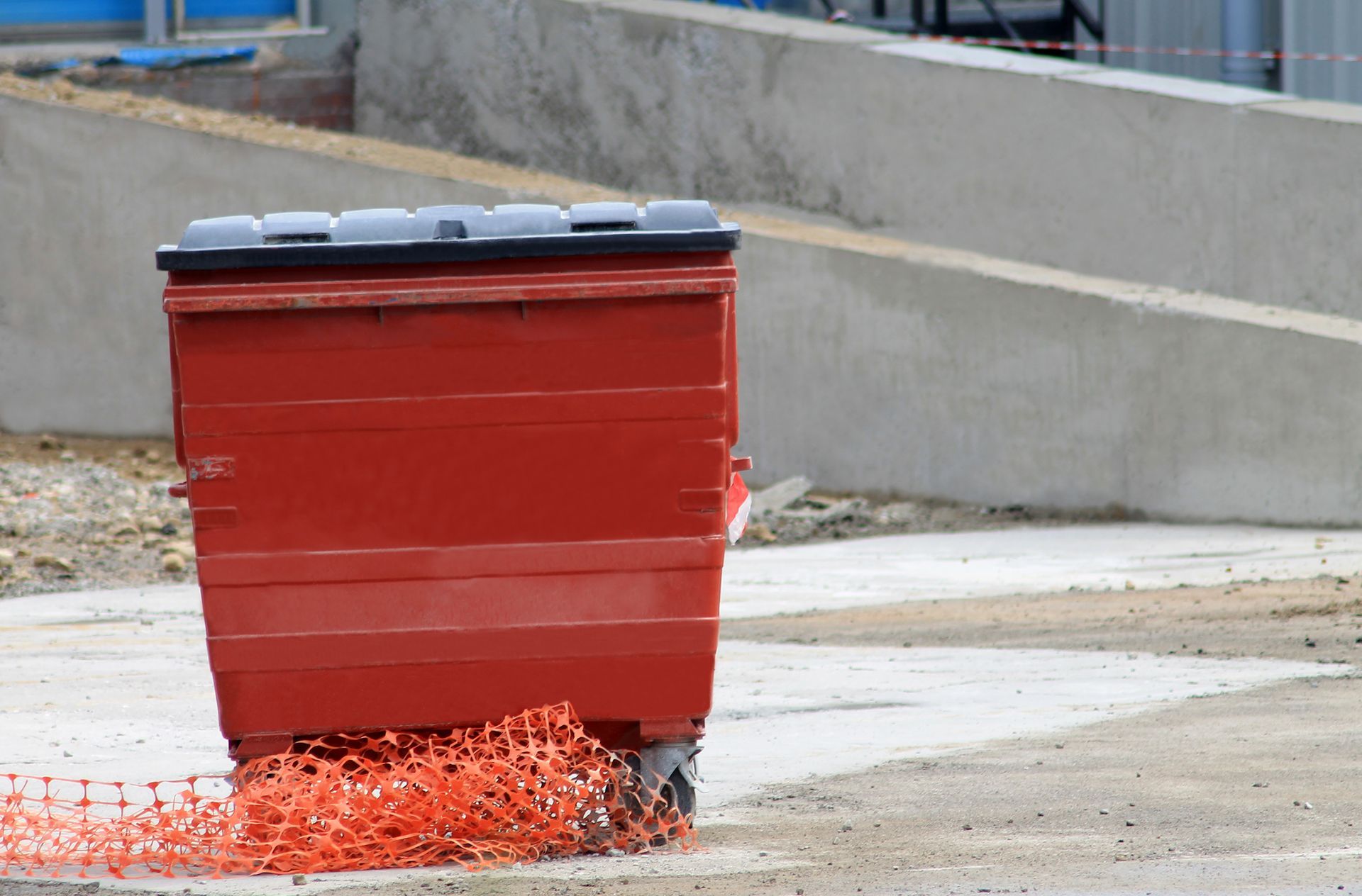 Red dumpster on wheels rolls over orange construction netting on a paved surface.