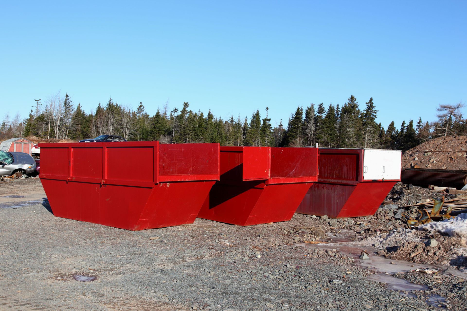 Three red dumpsters outdoors on a gravel lot under a clear blue sky.