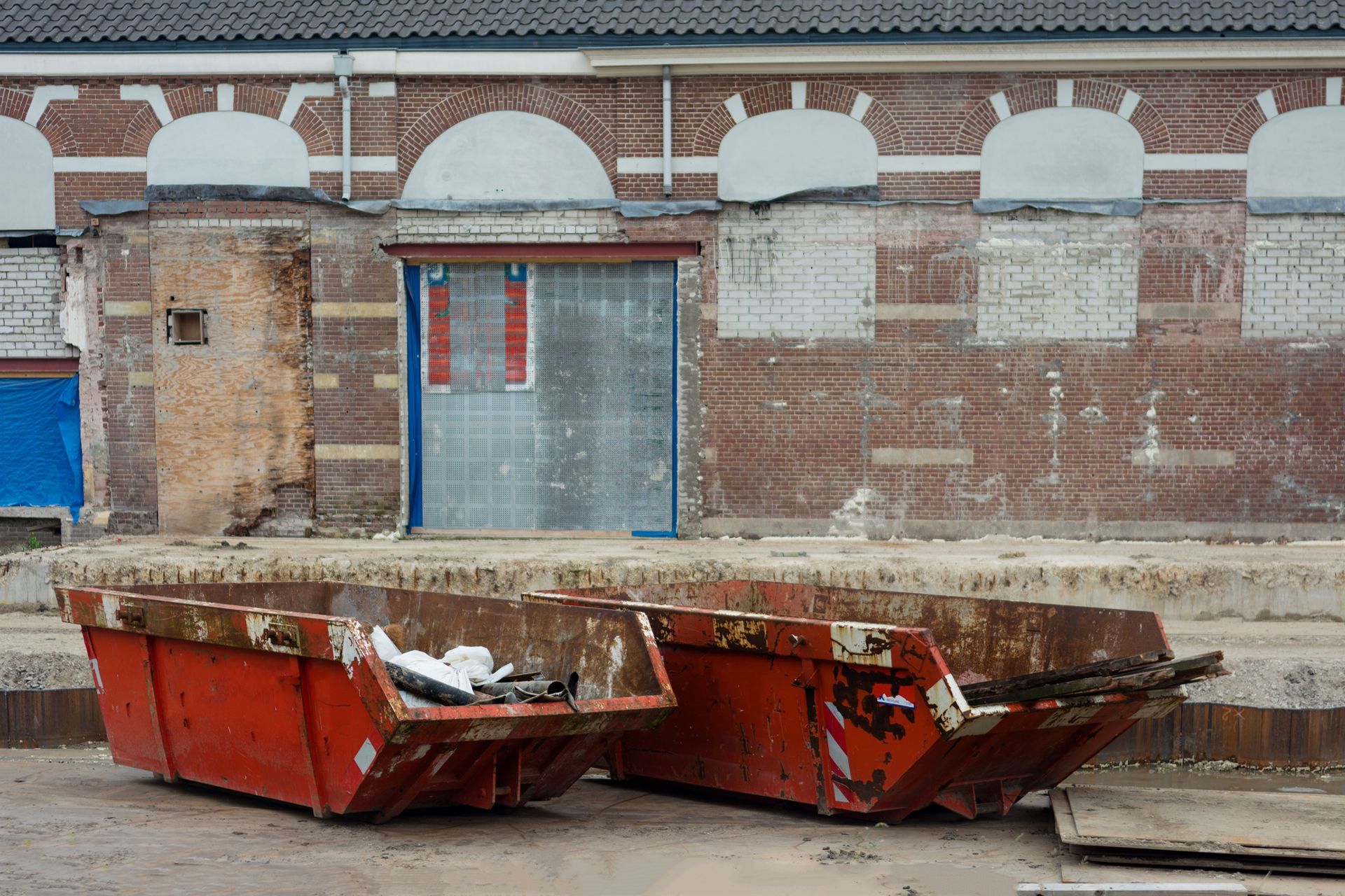 Two rusty red dumpsters in front of a partially demolished brick building with arched windows.