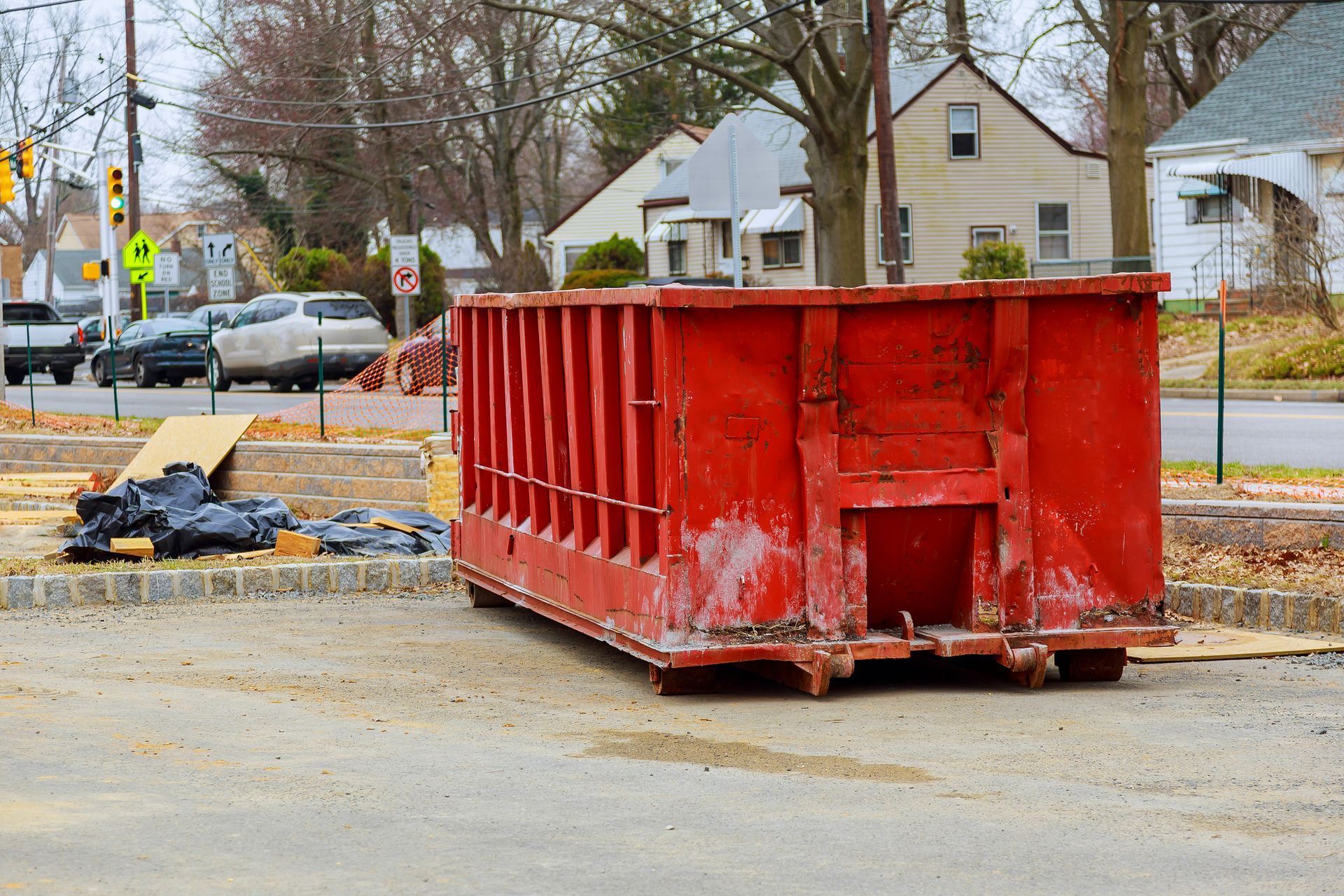 Red dumpster on a paved area, with debris beside it and houses in the background.