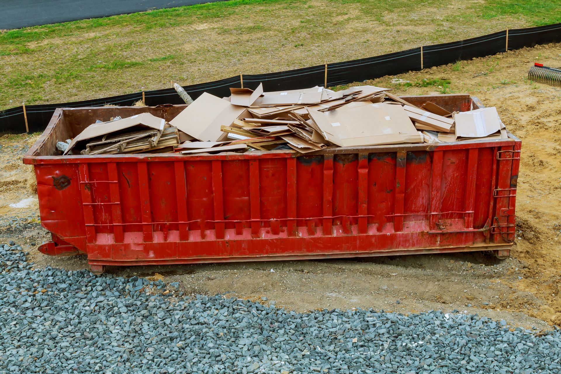 Red dumpster filled with cardboard debris, outdoors on gravel and dirt.