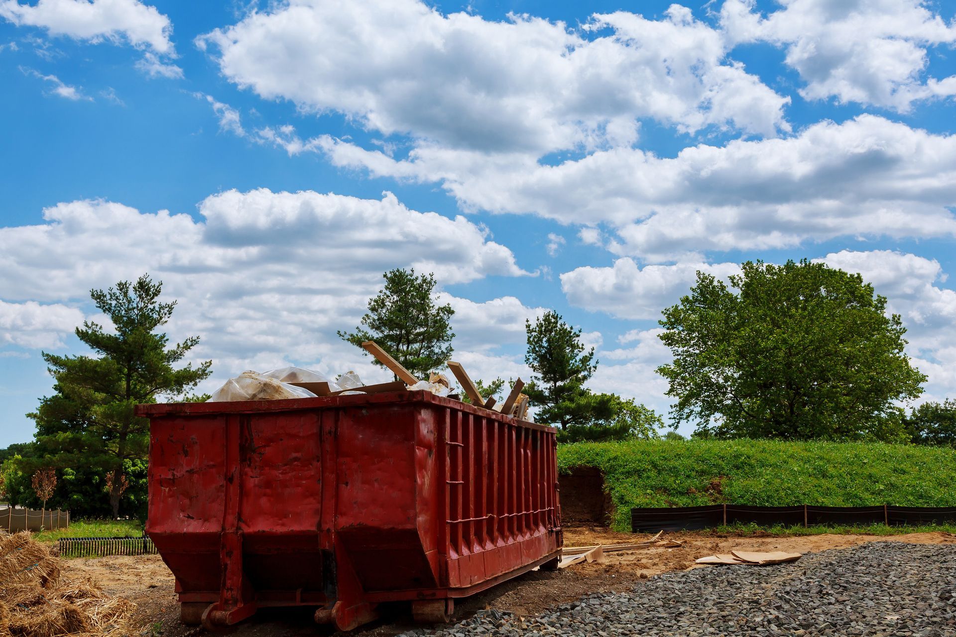 Red dumpster filled with debris on a construction site, under a cloudy blue sky.