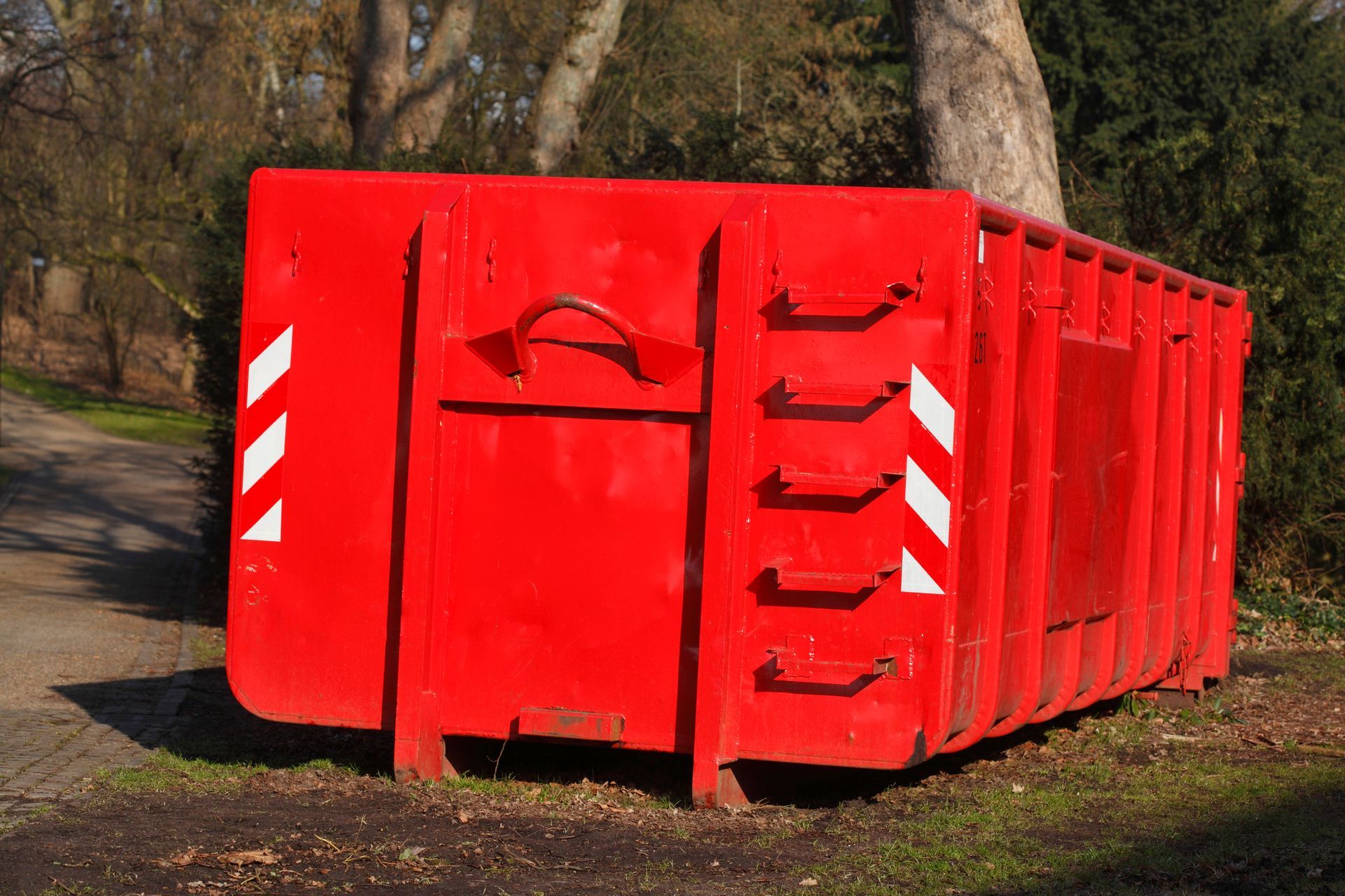 Red dumpster with white diagonal stripes, parked on grass near a path.