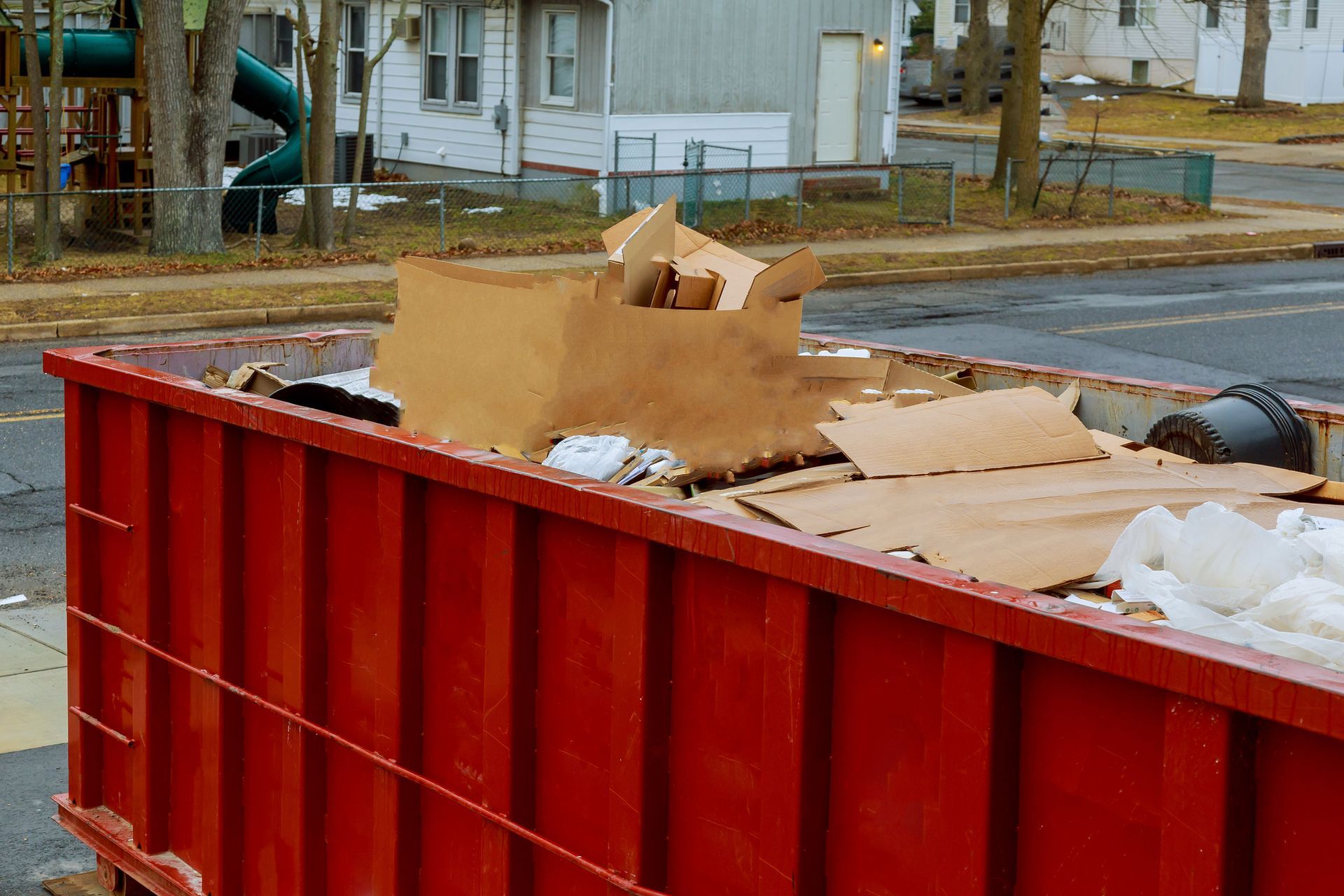Red dumpster filled with cardboard debris, outdoors near residential buildings and a playground.
