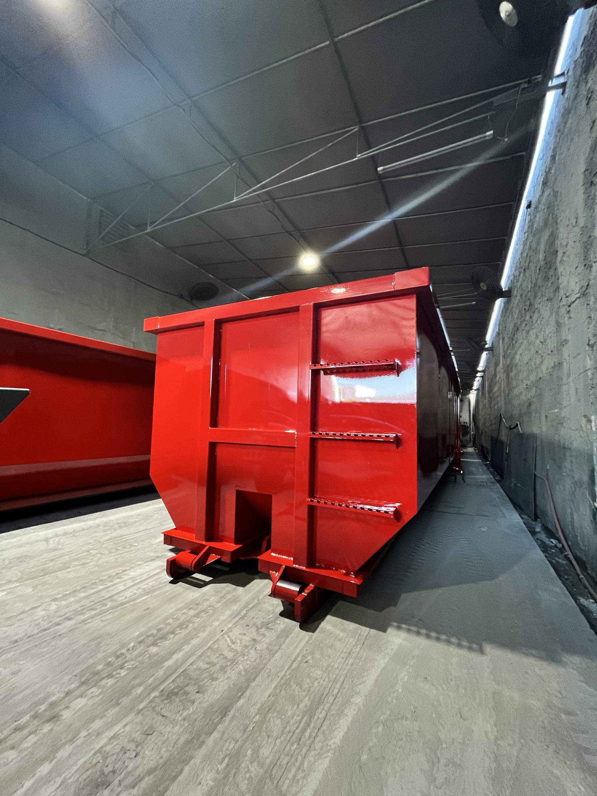 Red dumpster on a paved road, next to a white building and stone wall, under a clear blue sky.