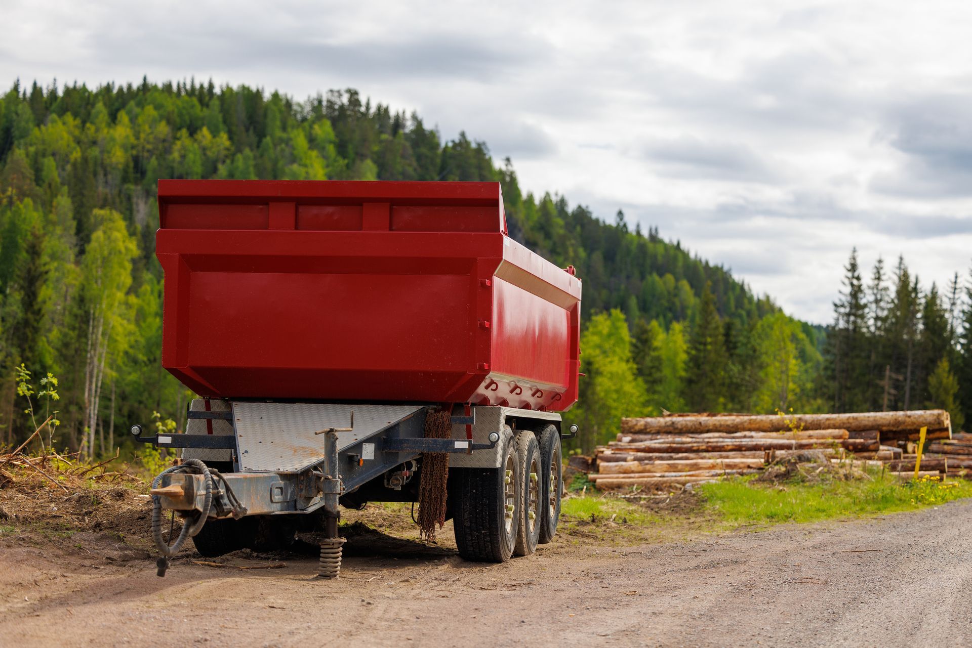 Red dump trailer parked on a dirt road in front of a forest with a pile of logs.