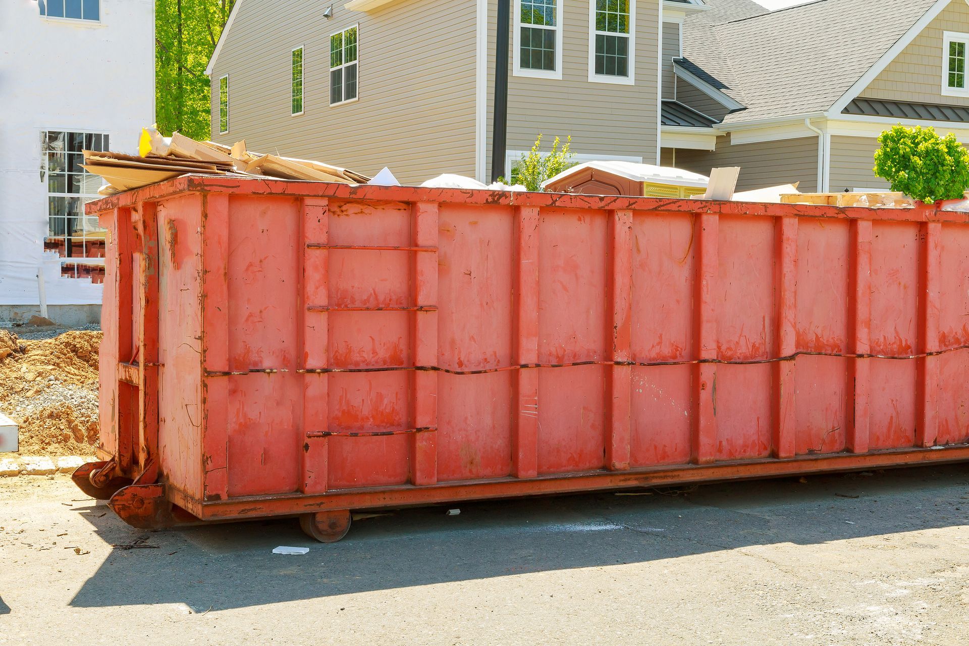 Red construction dumpster on a paved street, overflowing with debris, near new houses.