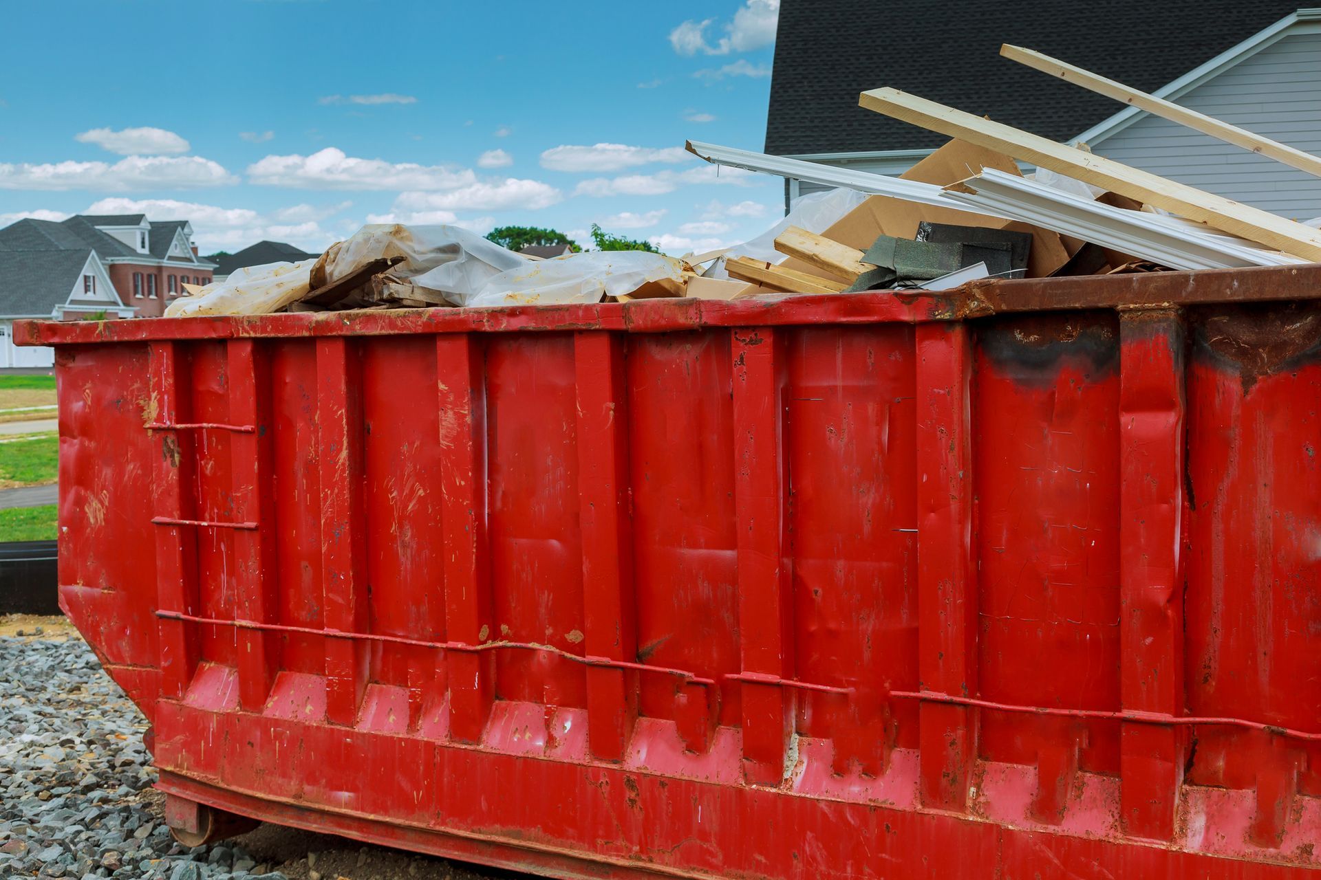 Red dumpster filled with construction debris, with houses and blue sky in the background.