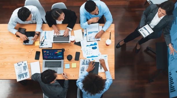 A group of people are sitting around a table with laptops and papers.