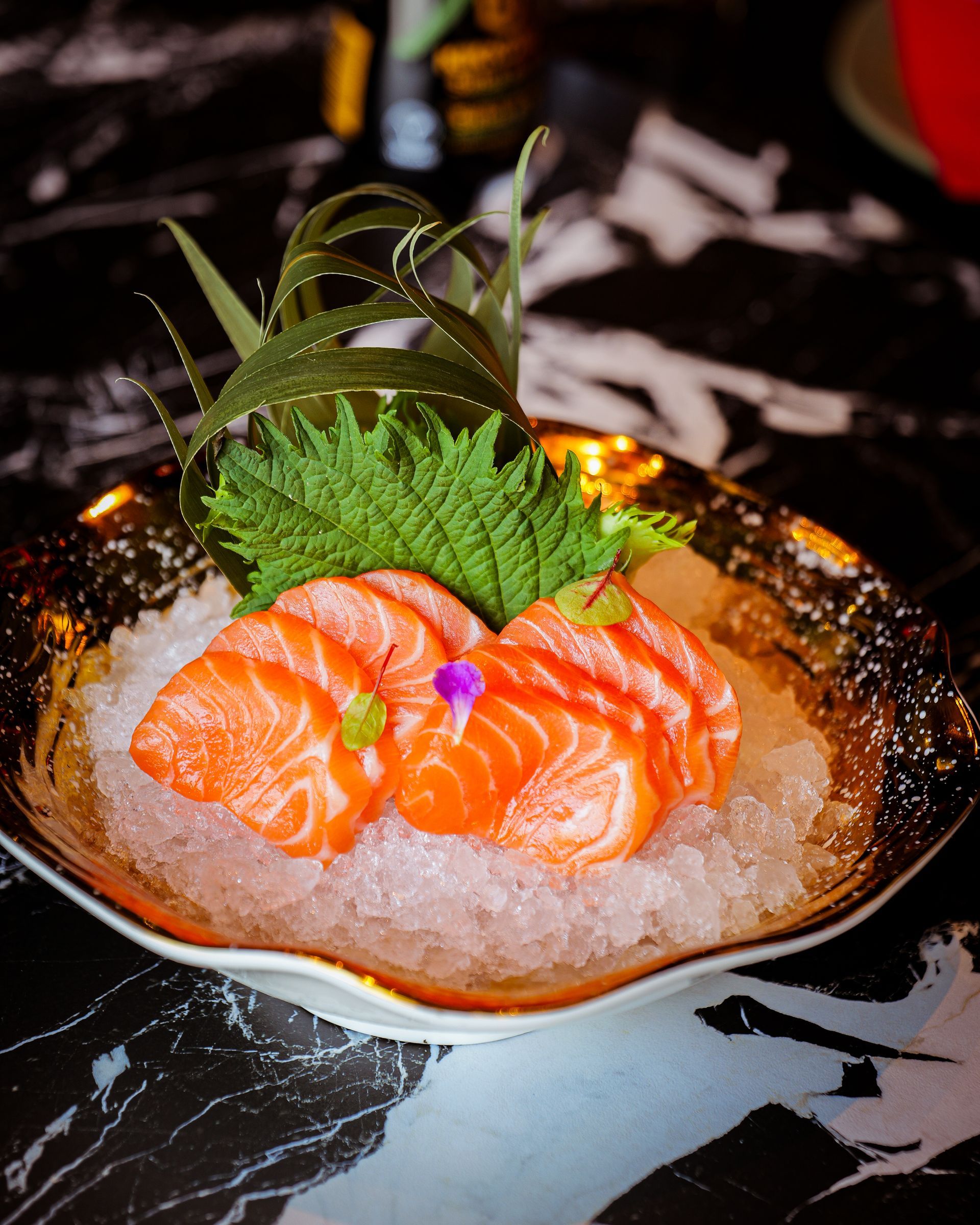 Salmone sashimi su ghiaccio tritato in una ciotola nera, guarnito con foglia di shiso e rametto di erbe aromatiche.