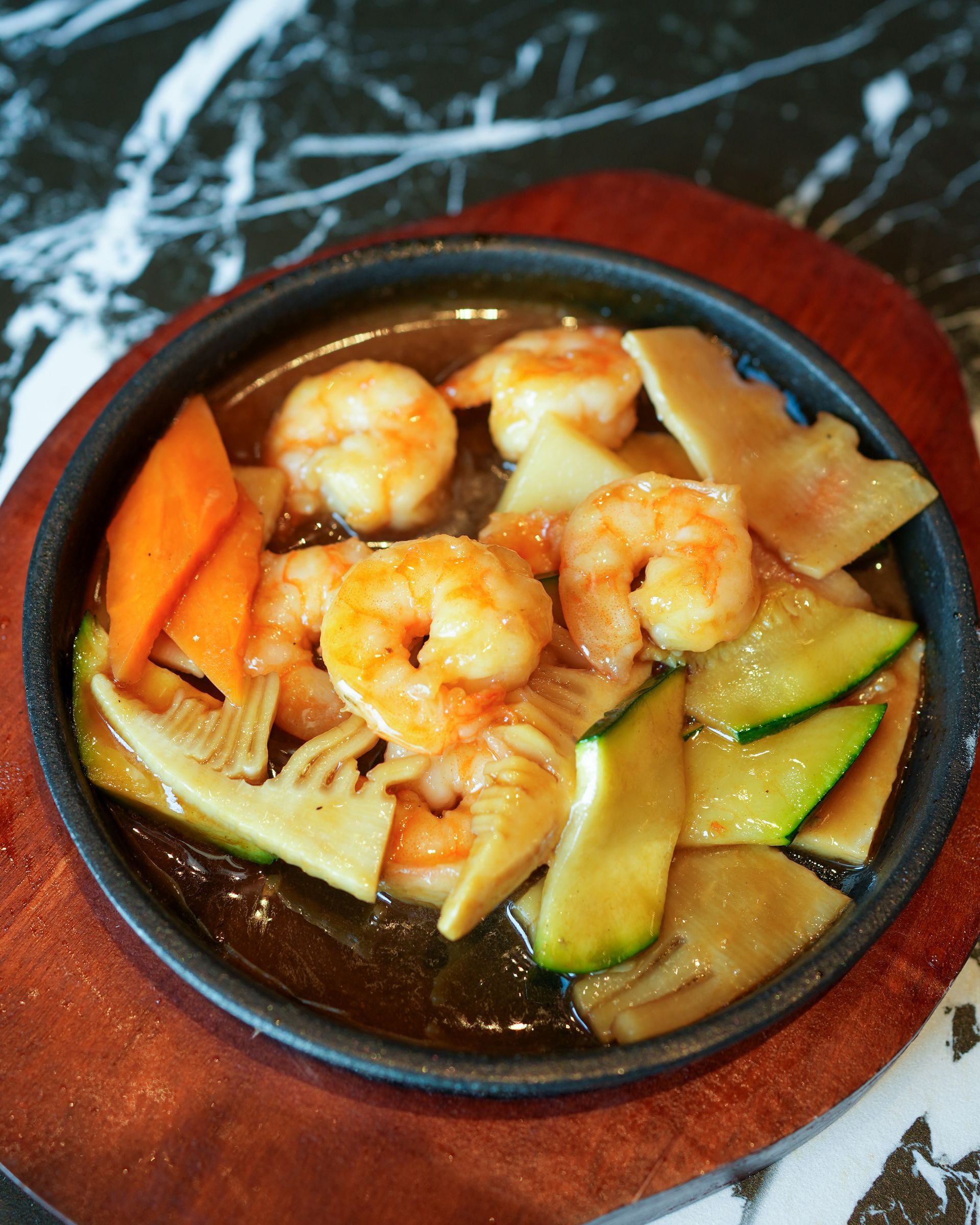 Gamberetti e verdure in una ciotola scura su un piatto di legno, con pezzi arancioni e verdi in brodo