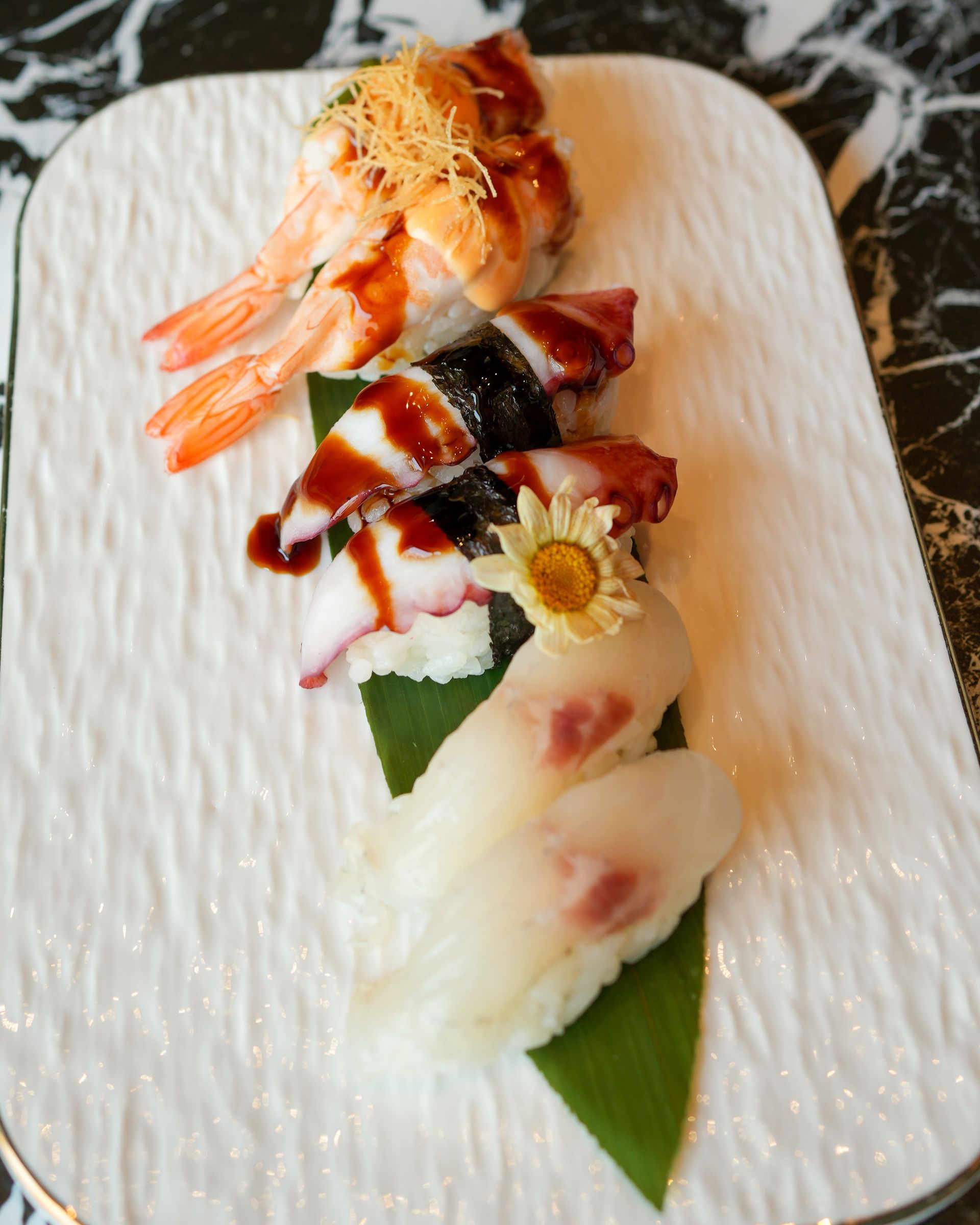 Assortimento di sushi su un piatto rettangolare bianco, guarnito con gamberi e salsa, su una foglia verde.