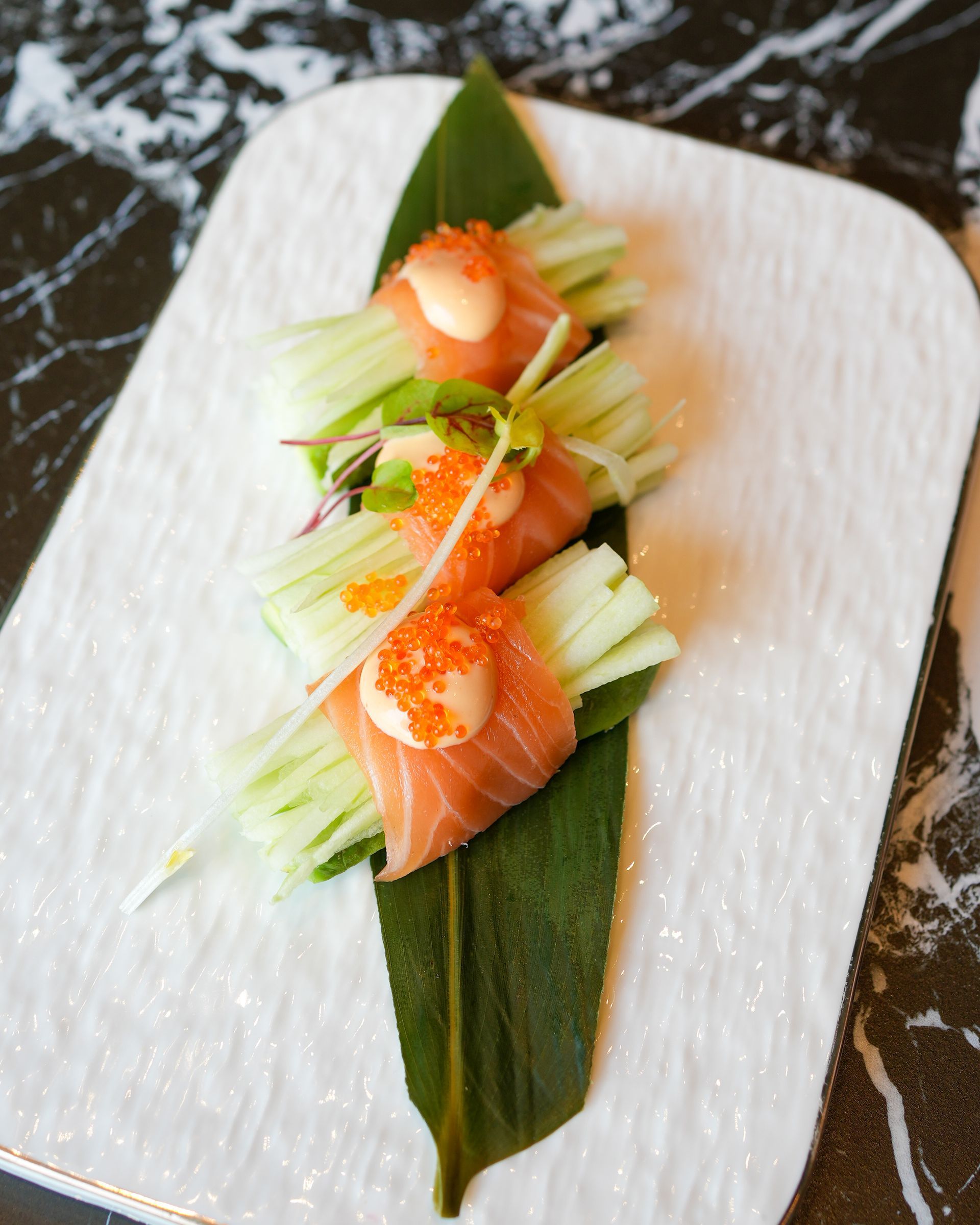 Tre nigiri di salmone guarniti con sottili fette di cetriolo su un piatto bianco dalla superficie ruvida, con una guarnizione di foglie verdi.
