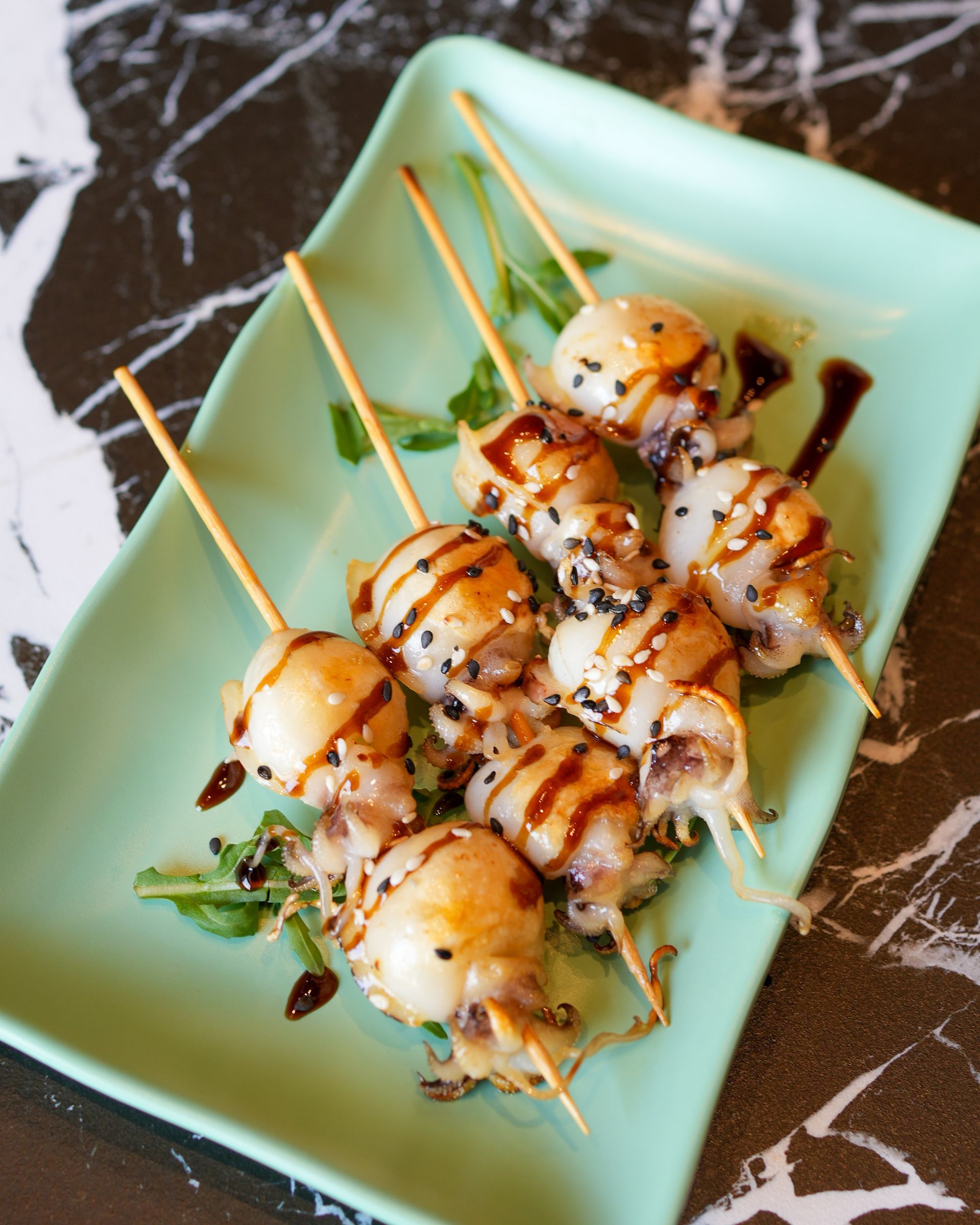 Capesante grigliate su spiedini, condite con salsa e servite su un piatto verde menta.