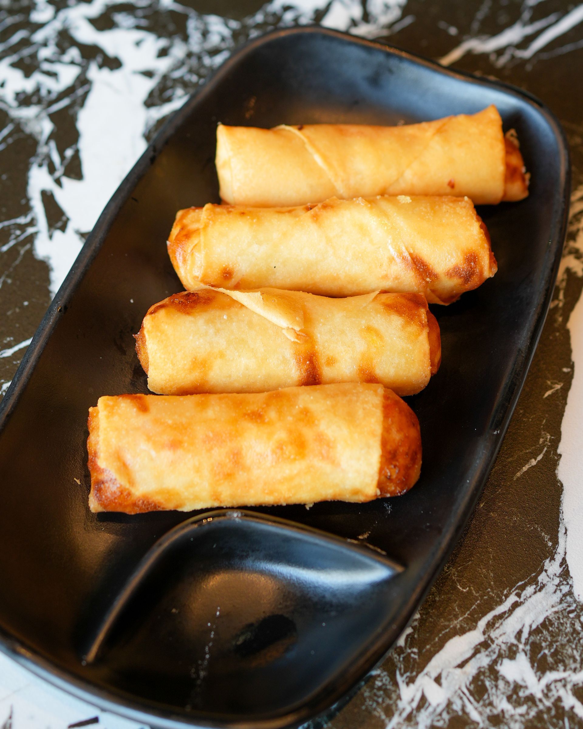 Quattro involtini primavera dorati su un piatto da portata nero con una piccola vaschetta per la salsa.