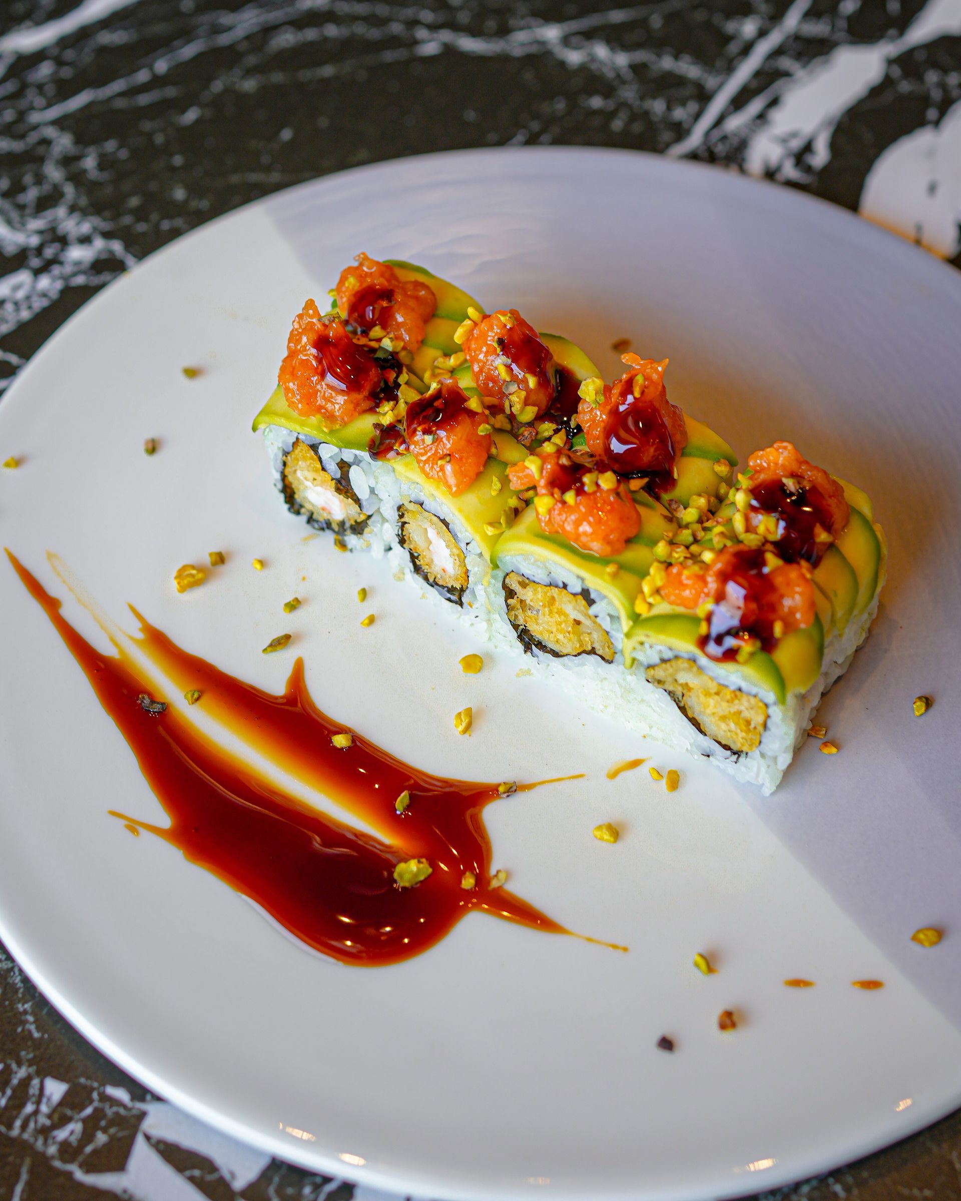 Rotolo di sushi guarnito con pesce a cubetti e salsa su un piatto bianco con una spruzzata di salsa rossa.