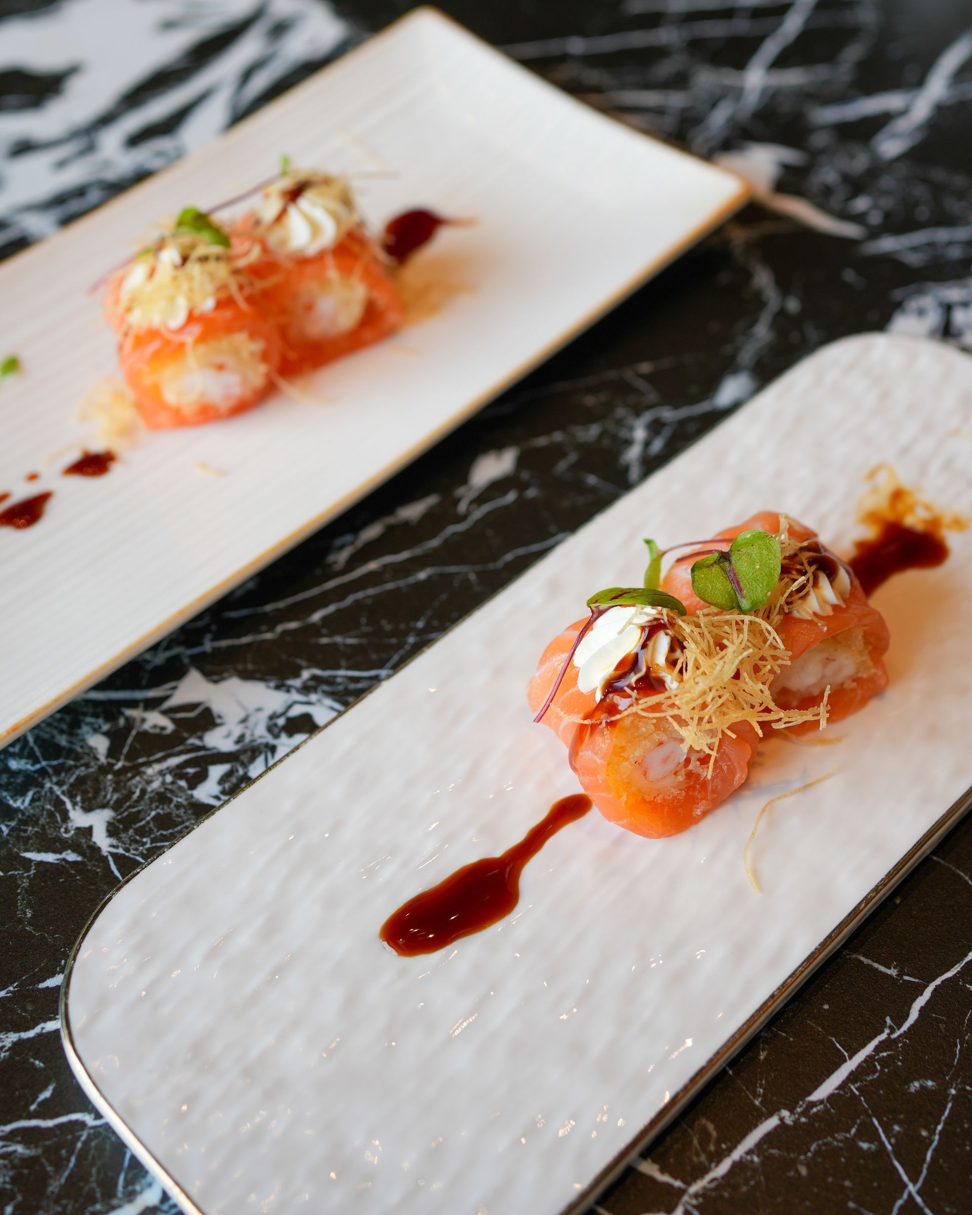 Due involtini di sushi serviti su piatti rettangolari bianchi con salsa scura, su un tavolo di marmo nero.