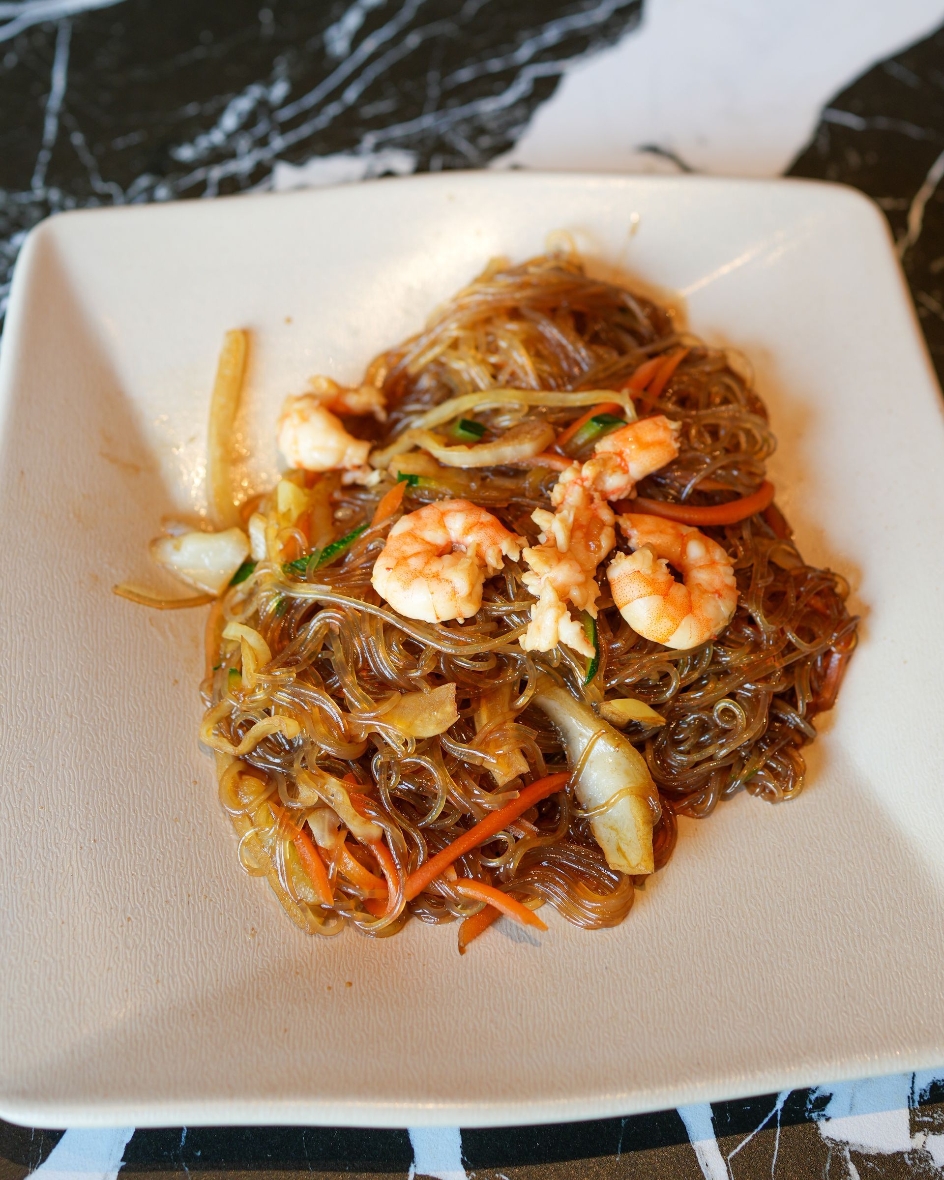 Saltato in padella con gamberi, noodles e verdure, servito su un piatto quadrato bianco.