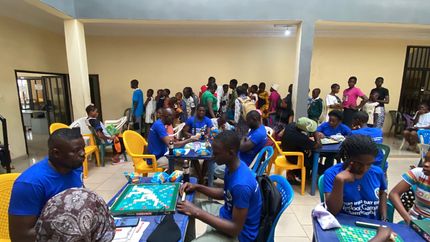 People seated at tables playing a game, some in blue shirts, in a brightly lit room with a crowd of people in the background.