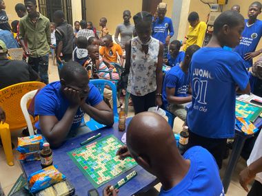 People playing board games in a crowded room. Two players at a table, others watching. Blue and yellow clothing.