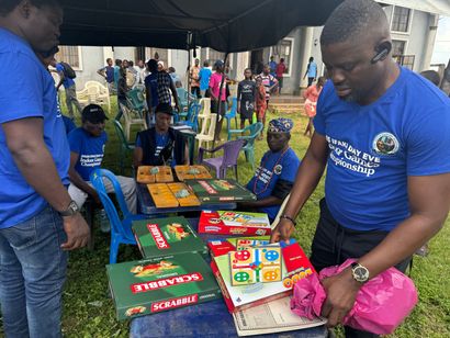 People at a table with board games, under a tent outdoors. One man looks at a board game. Others seated.