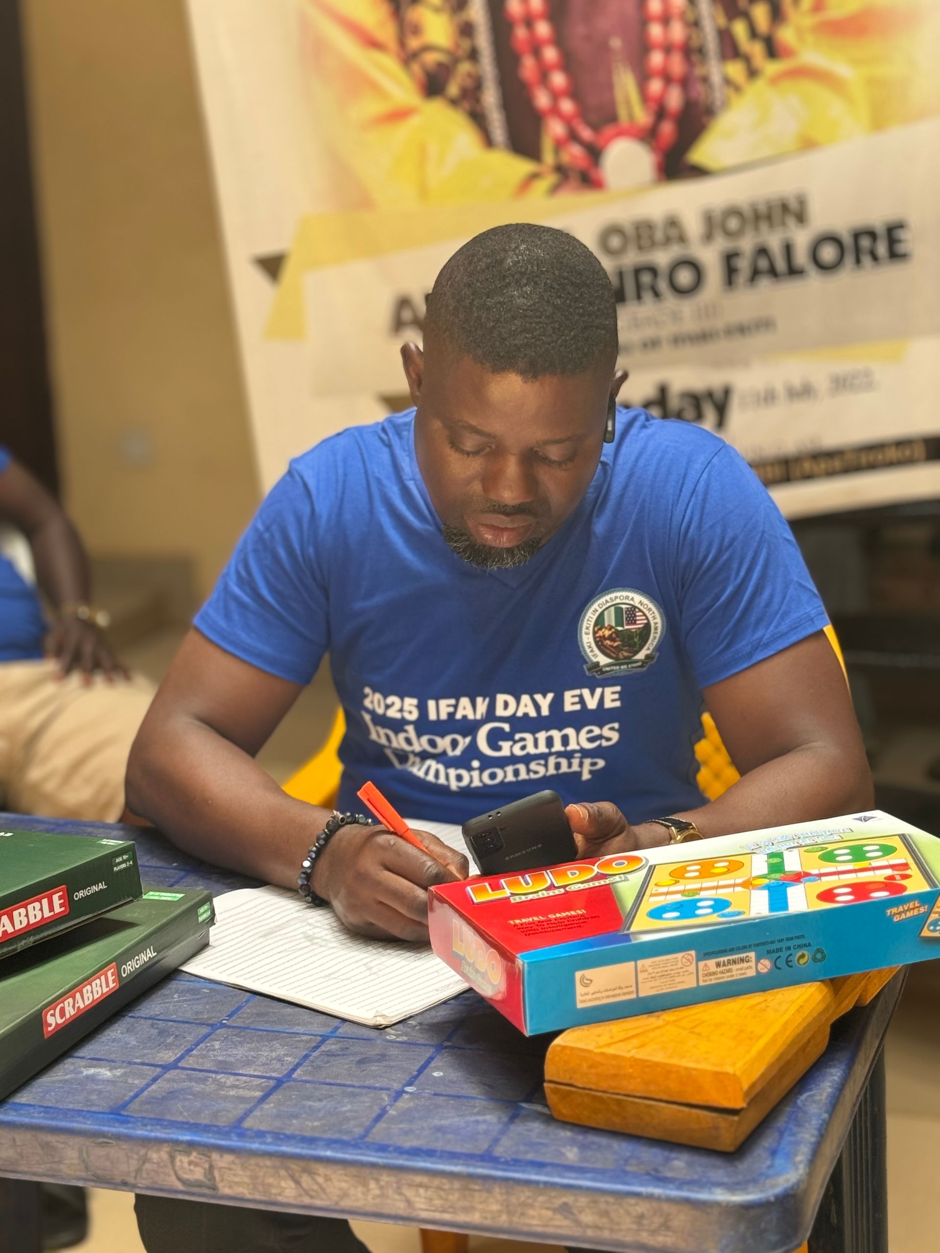 Man in blue shirt writing at a table, game boards present, poster in background.