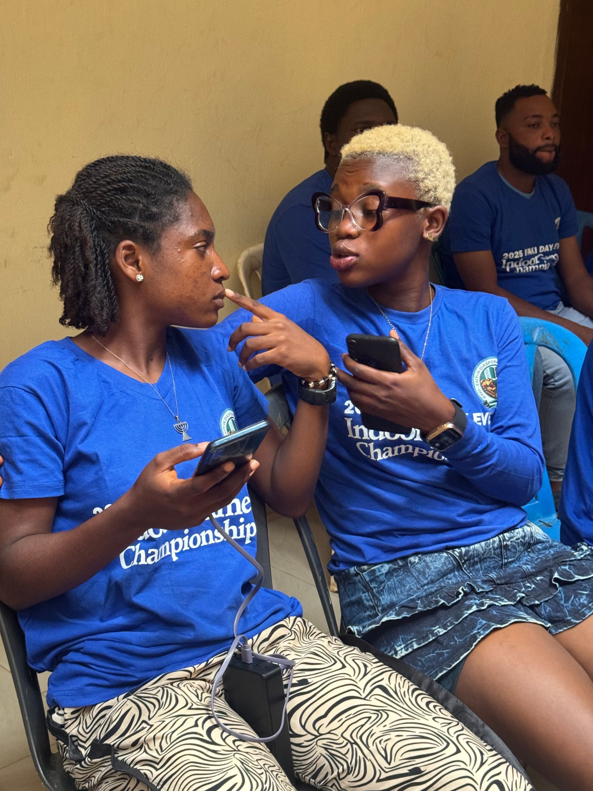 Two people in blue shirts are talking. One points at the other while holding a phone. Others in the background.