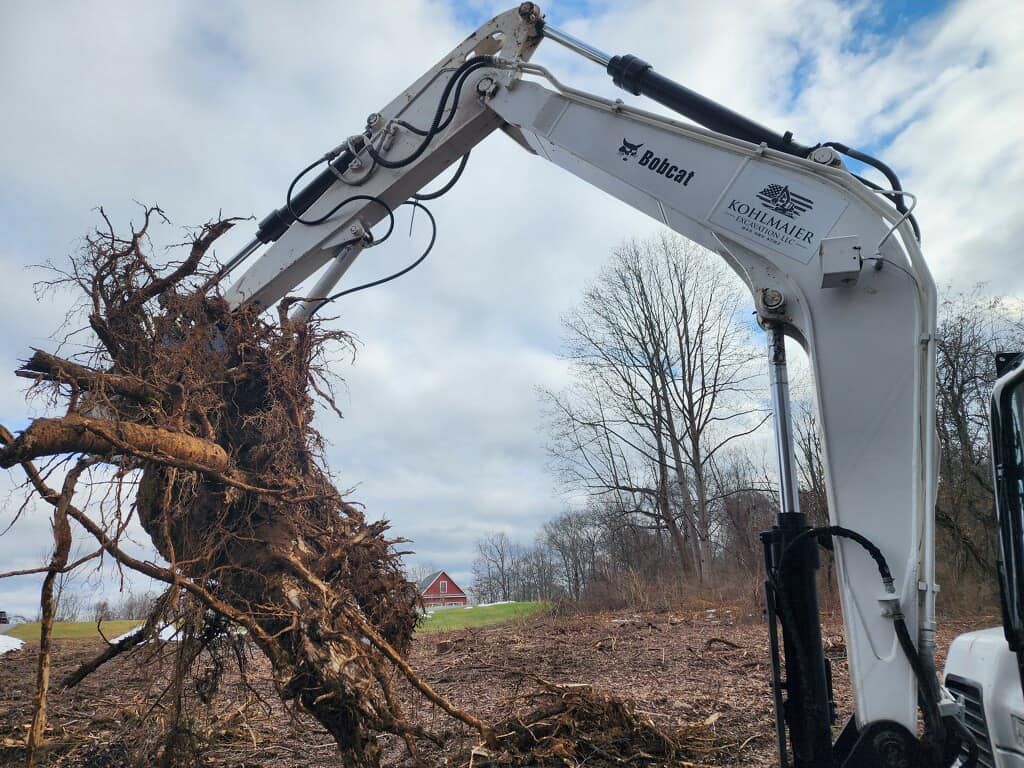 A white excavator is carrying a large tree stump in its bucket.
