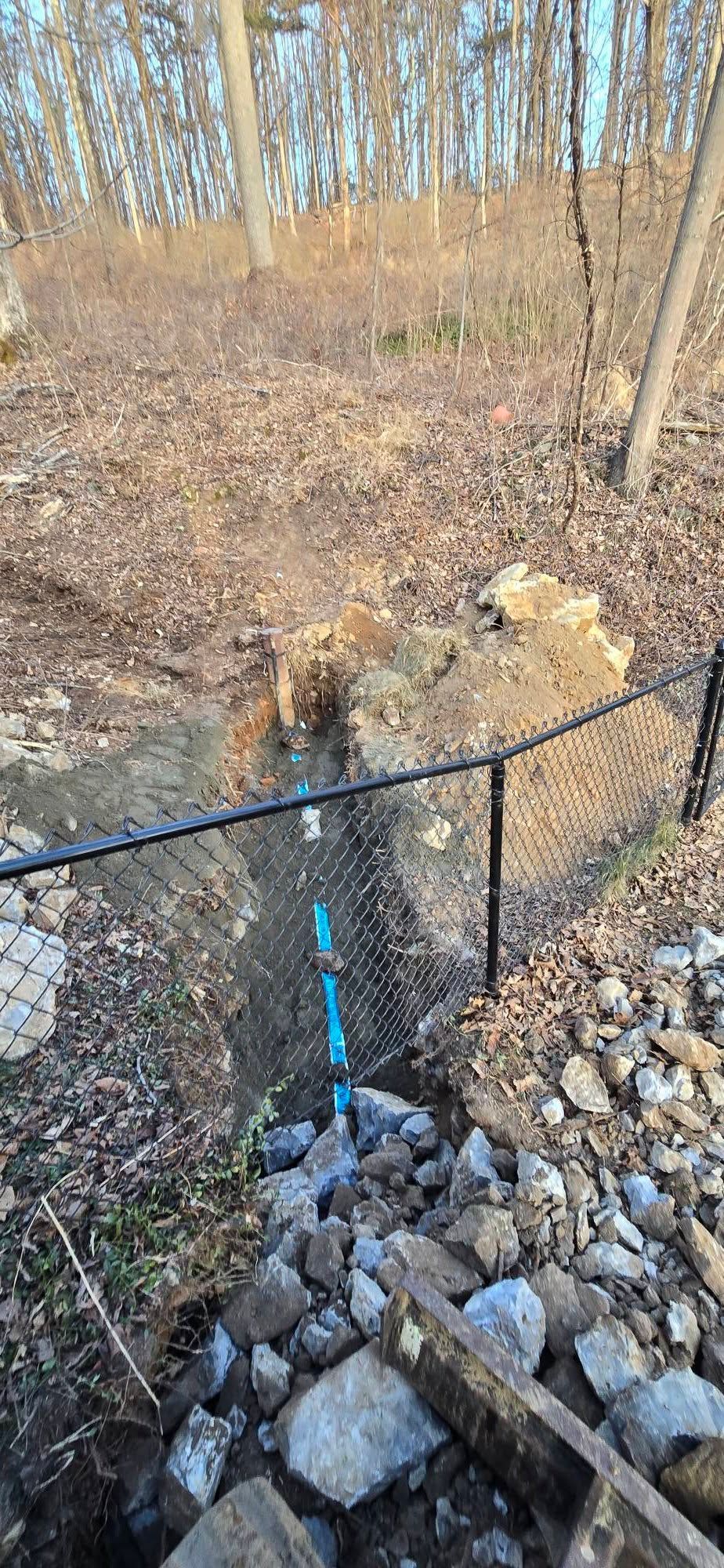 A blue pipe is going through a chain link fence in the woods.