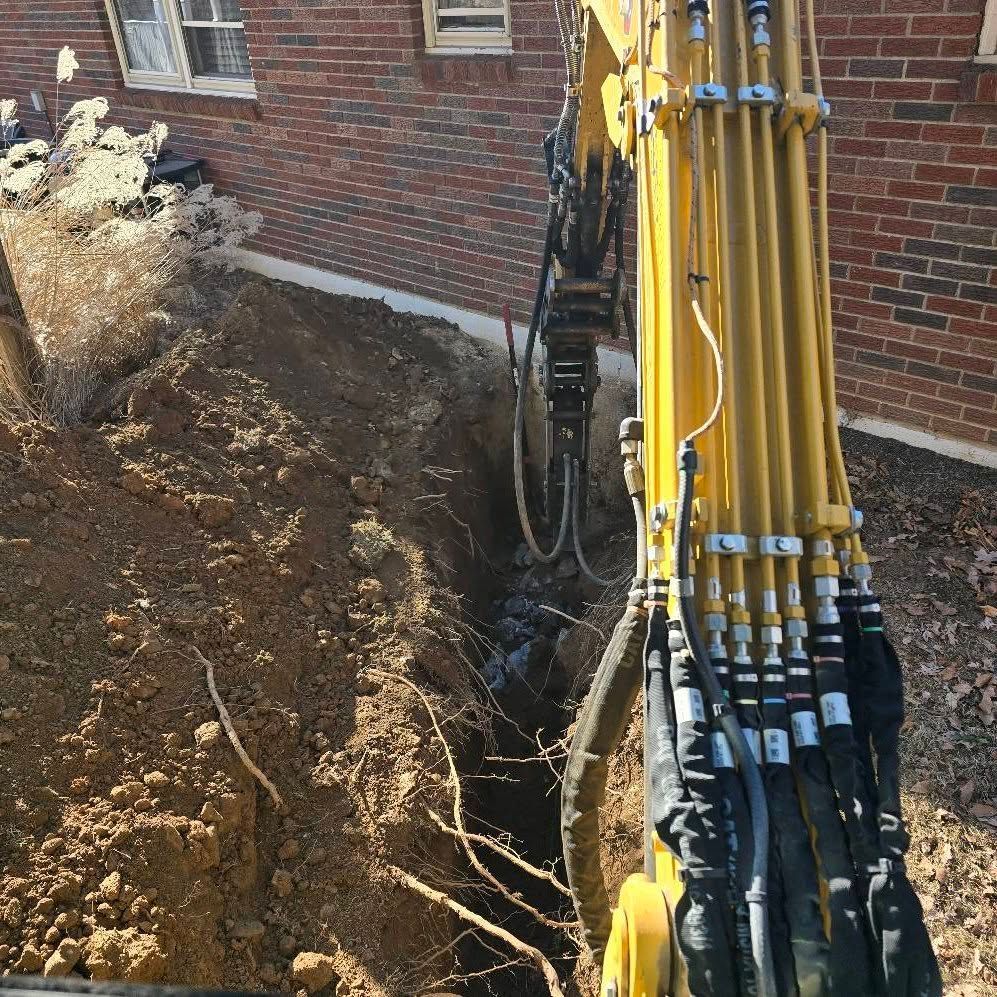 A yellow excavator is digging a hole in the ground in front of a brick building.