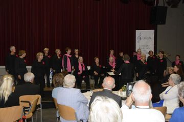 Een koor en dirigent treden op op het podium, met een publiek vooraan. De koorleden dragen formele kleding.