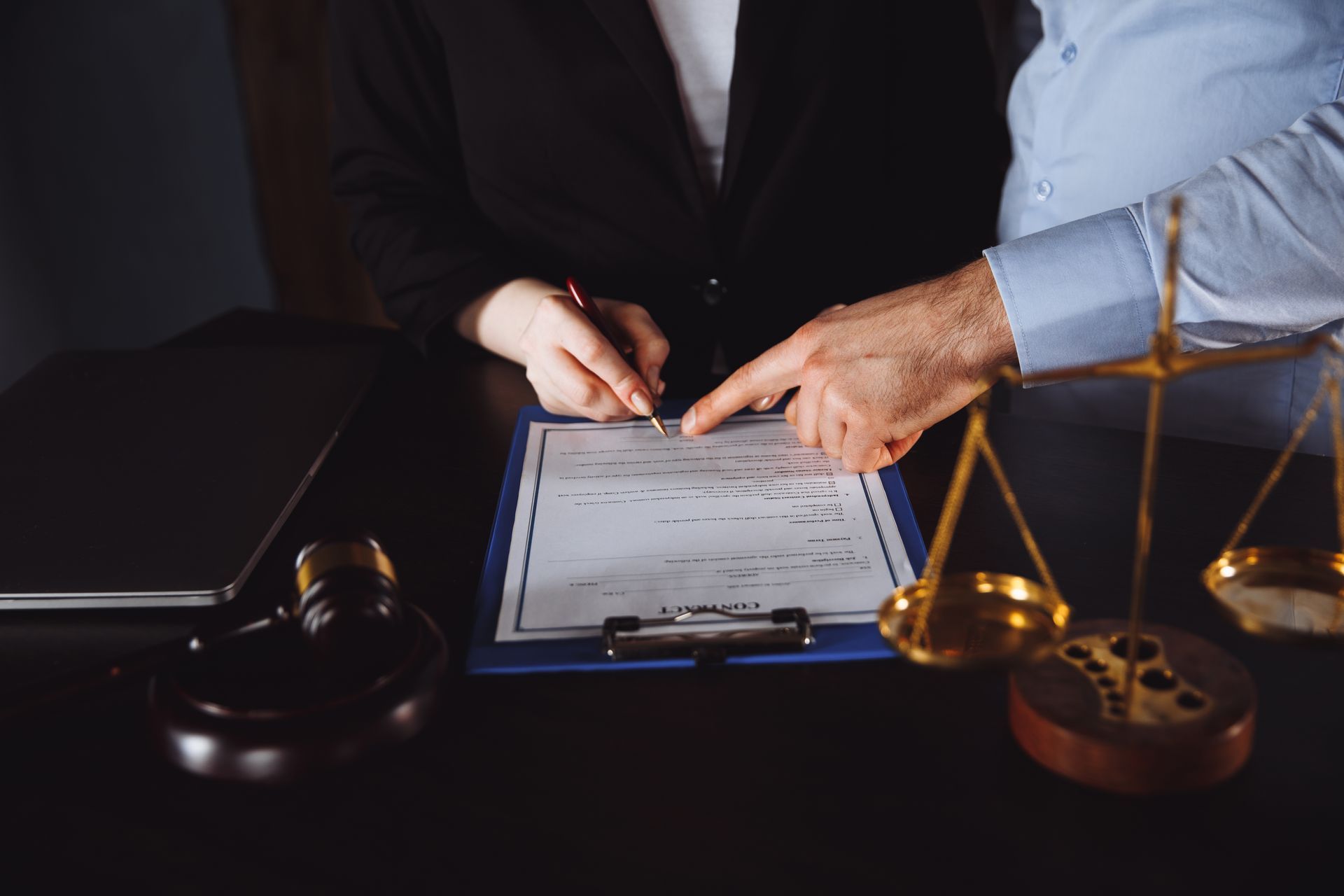 Person signing legal document; scales of justice, gavel, and laptop on table.