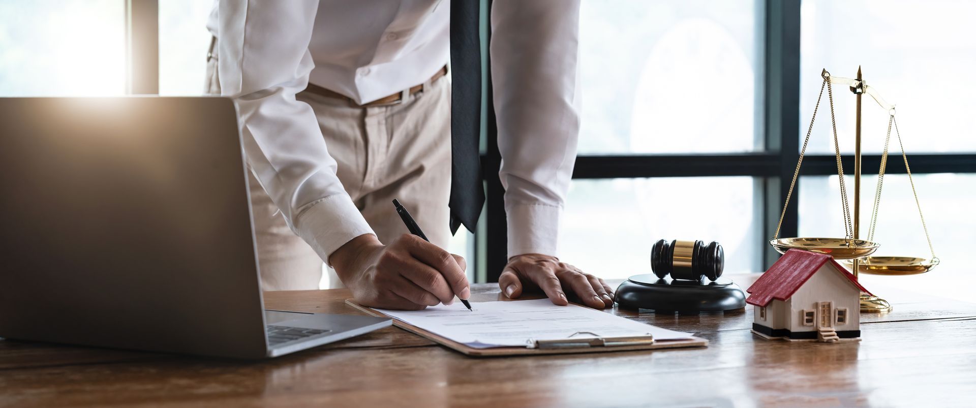 Person signing legal documents on a desk with a laptop, gavel, scales, and a miniature house.