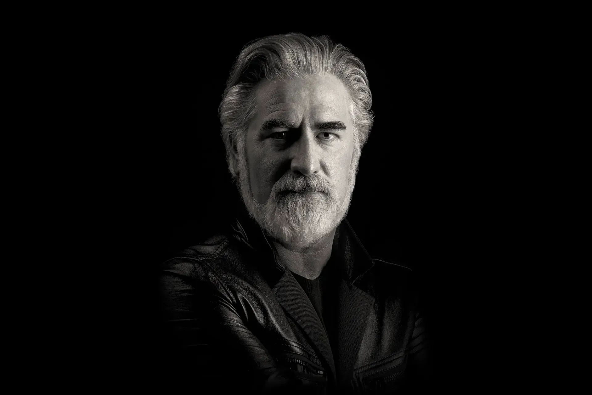 Man with gray hair and beard, wearing a black jacket, looking directly at the viewer against a black background.